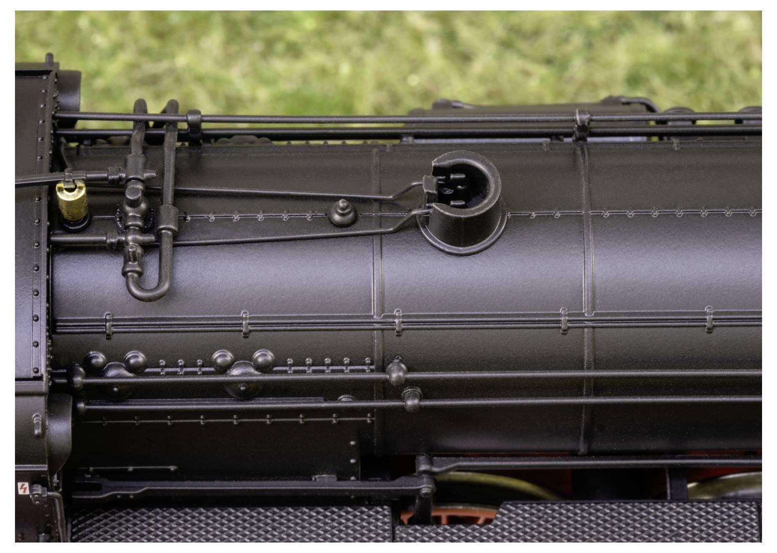 Nahaufnahme eines schwarzen Lokomotivkessels mit detaillierter Metallarbeit und Rohrleitungen vor einem unscharfen grünen Hintergrund.