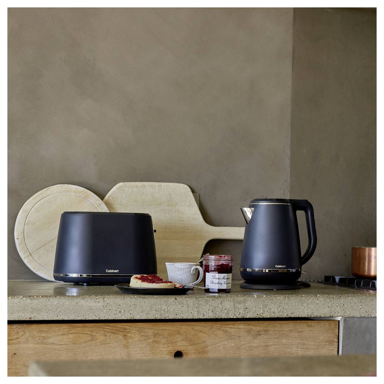 Eine Küchentheke zeigt einen schwarzen Toaster und Wasserkocher, eine Scheibe Brot mit Marmelade, ein Marmeladenglas und eine weiße Tasse, mit Schneidebrettern im Hintergrund.