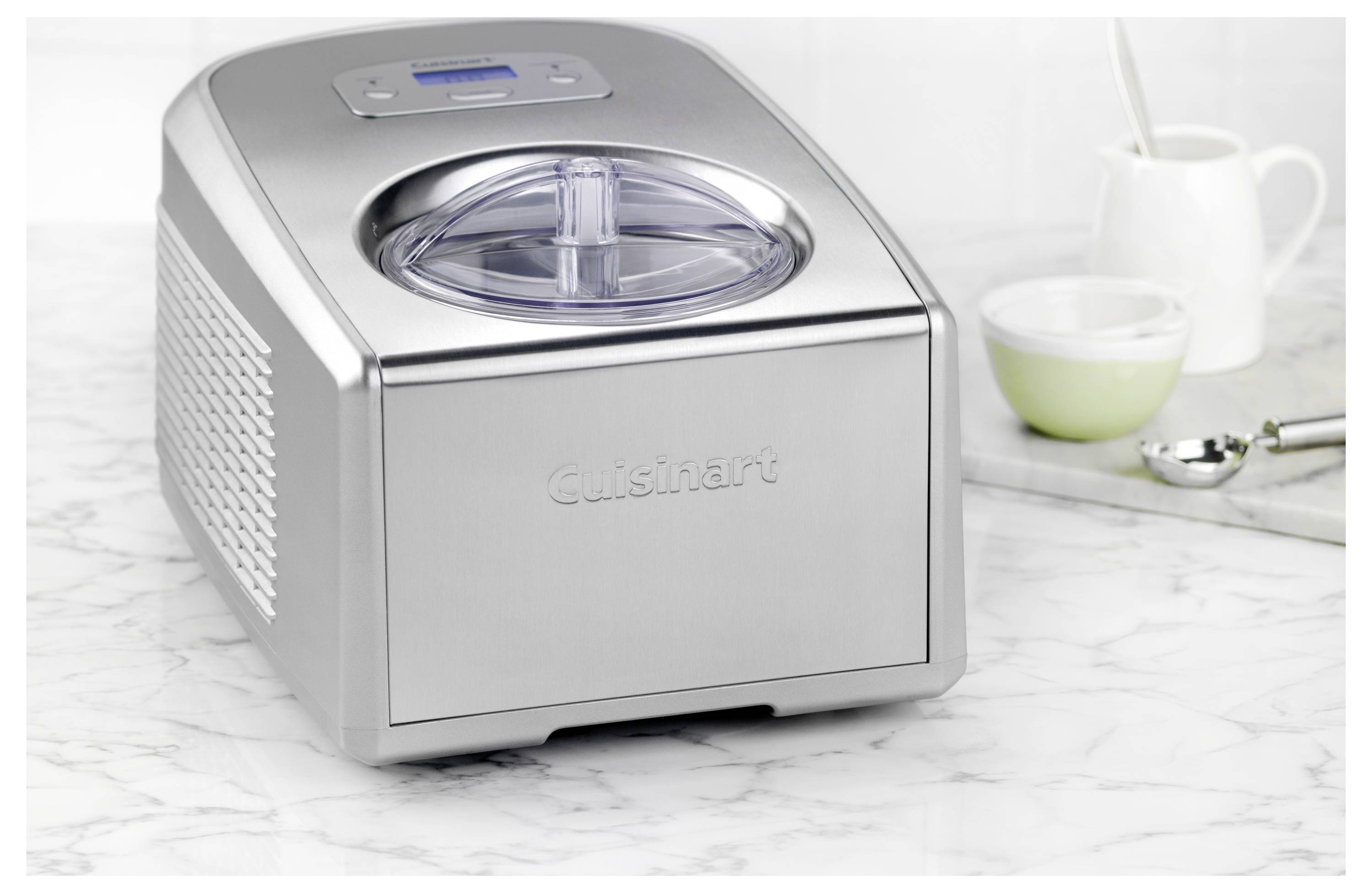 Eine Eismaschine aus Edelstahl auf einer Küchenarbeitsplatte, neben einer grünen Schüssel, einem Löffel und einem weißen Milchkännchen.