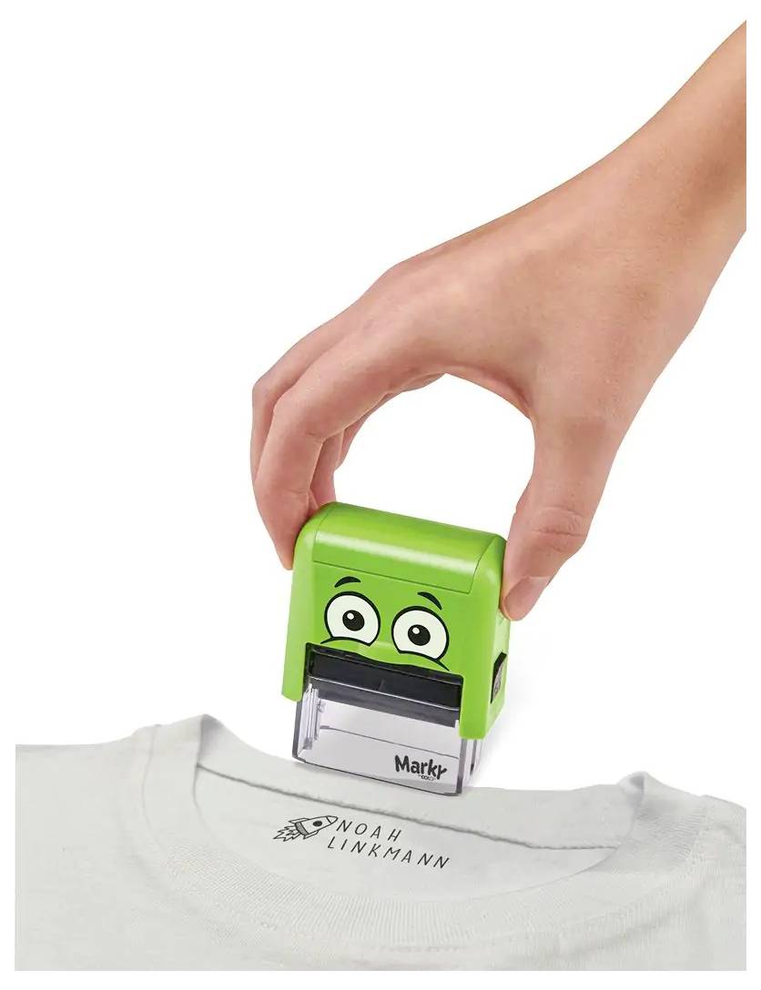 Eine Hand stempelt ein Etikett mit einem grünen Zeichen auf ein weißes Hemd, wobei nahe dem Kragen 'NOAH LINKMANN' gedruckt ist.