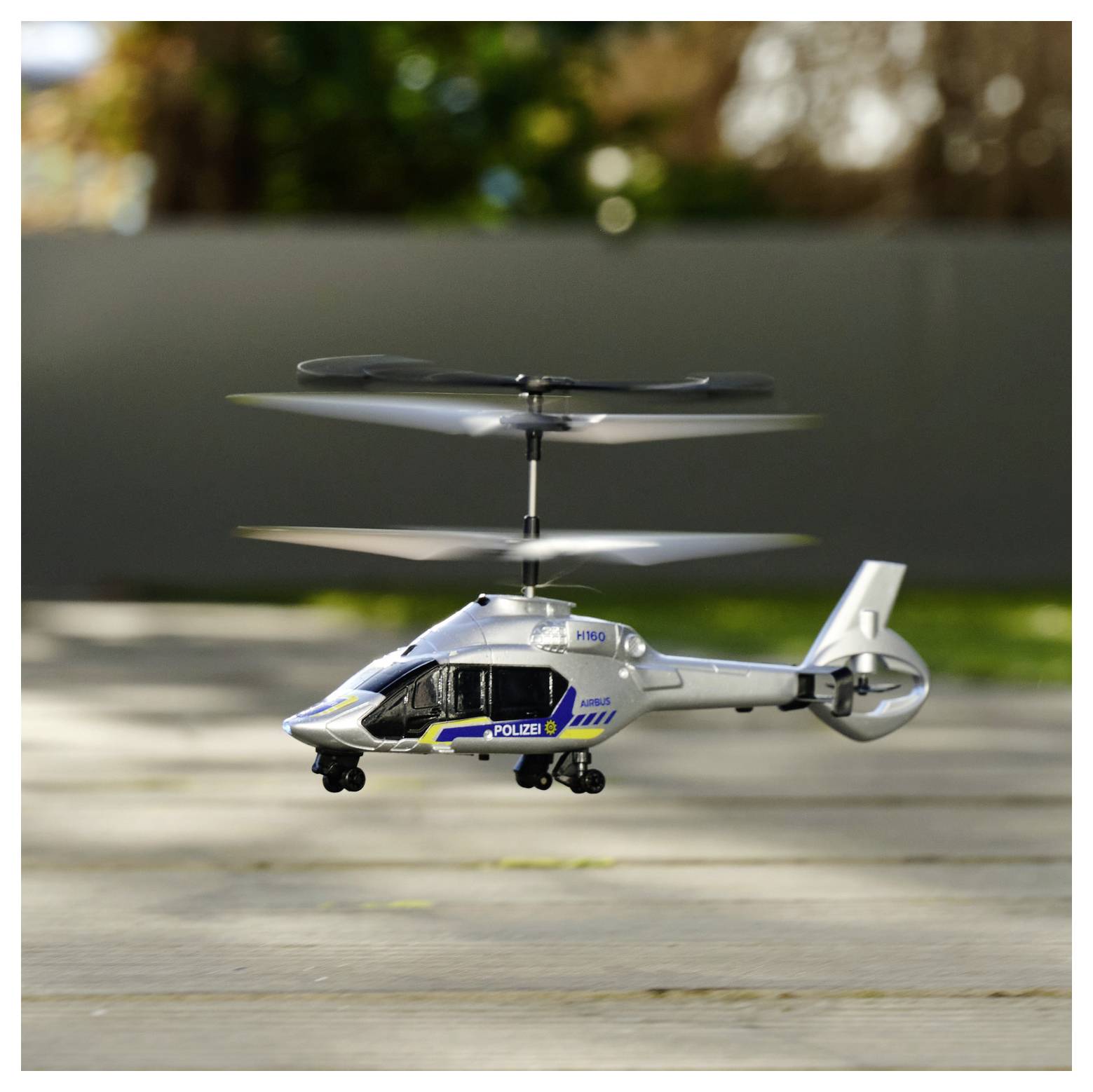 Ein kleiner Spielzeug-Hubschrauber mit der Aufschrift "Polizei" schwebt nahe dem Boden in einer Außenumgebung, während im Hintergrund Grünpflanzen unscharf zu sehen sind.