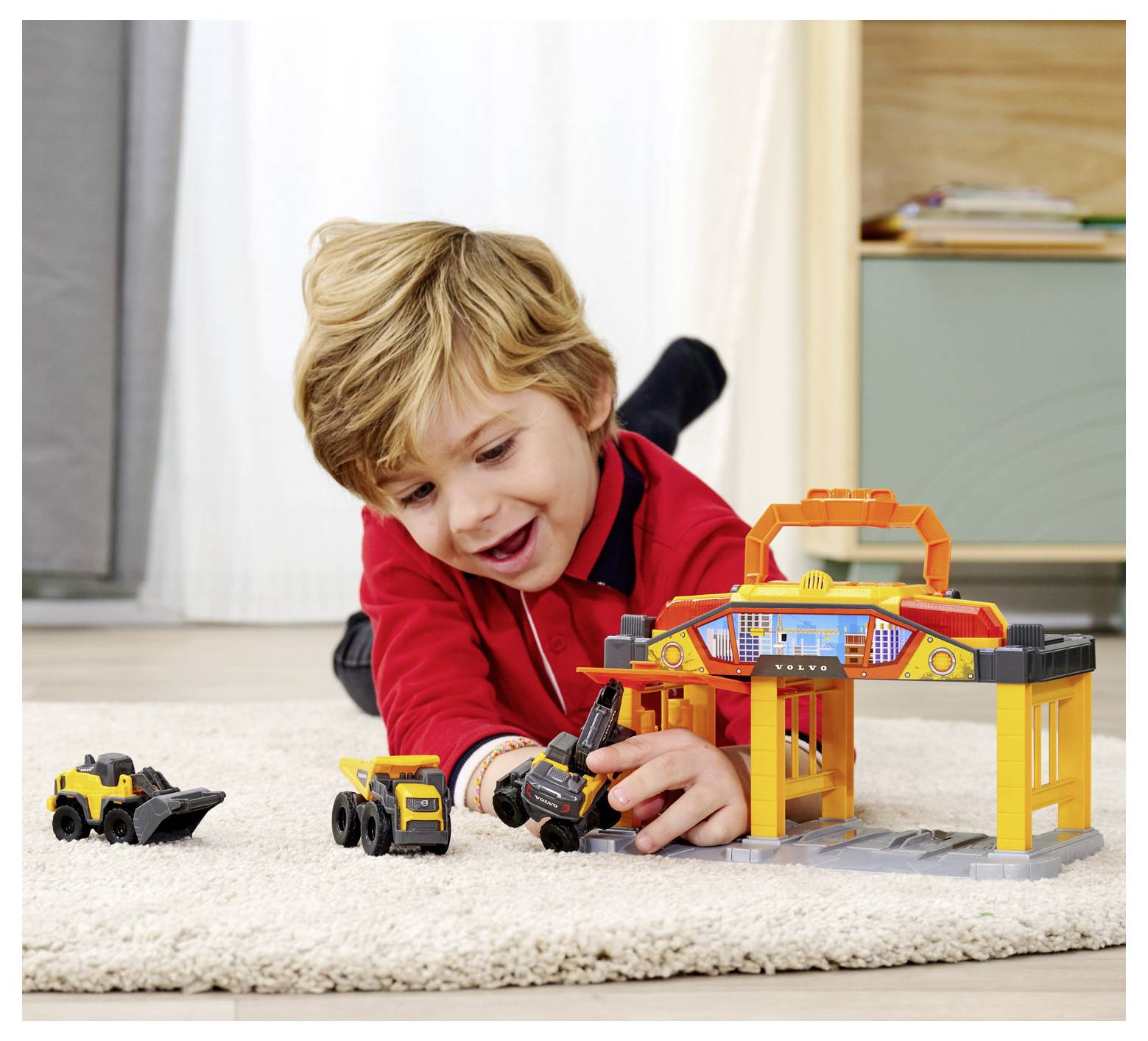 Ein Kind in einem roten Shirt spielt auf einem Teppich mit Spielzeug-Baufahrzeugen und einer Spielzeug-Garage. Die Umgebung ist eine gemütliche Inneneinrichtung.