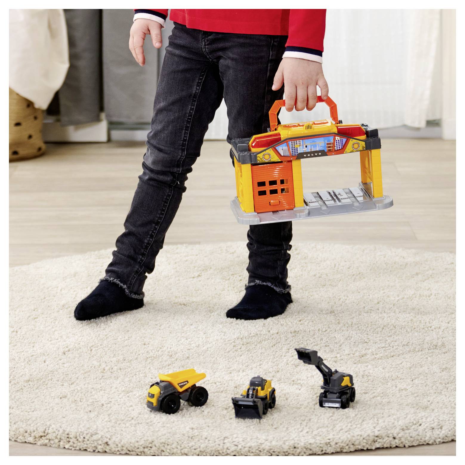 Ein Kind, das eine Spielzeuggarage hält, steht auf einem Teppich neben drei kleinen Baufahrzeugen und deutet damit auf phantasievolles Spielen zu Hause hin.