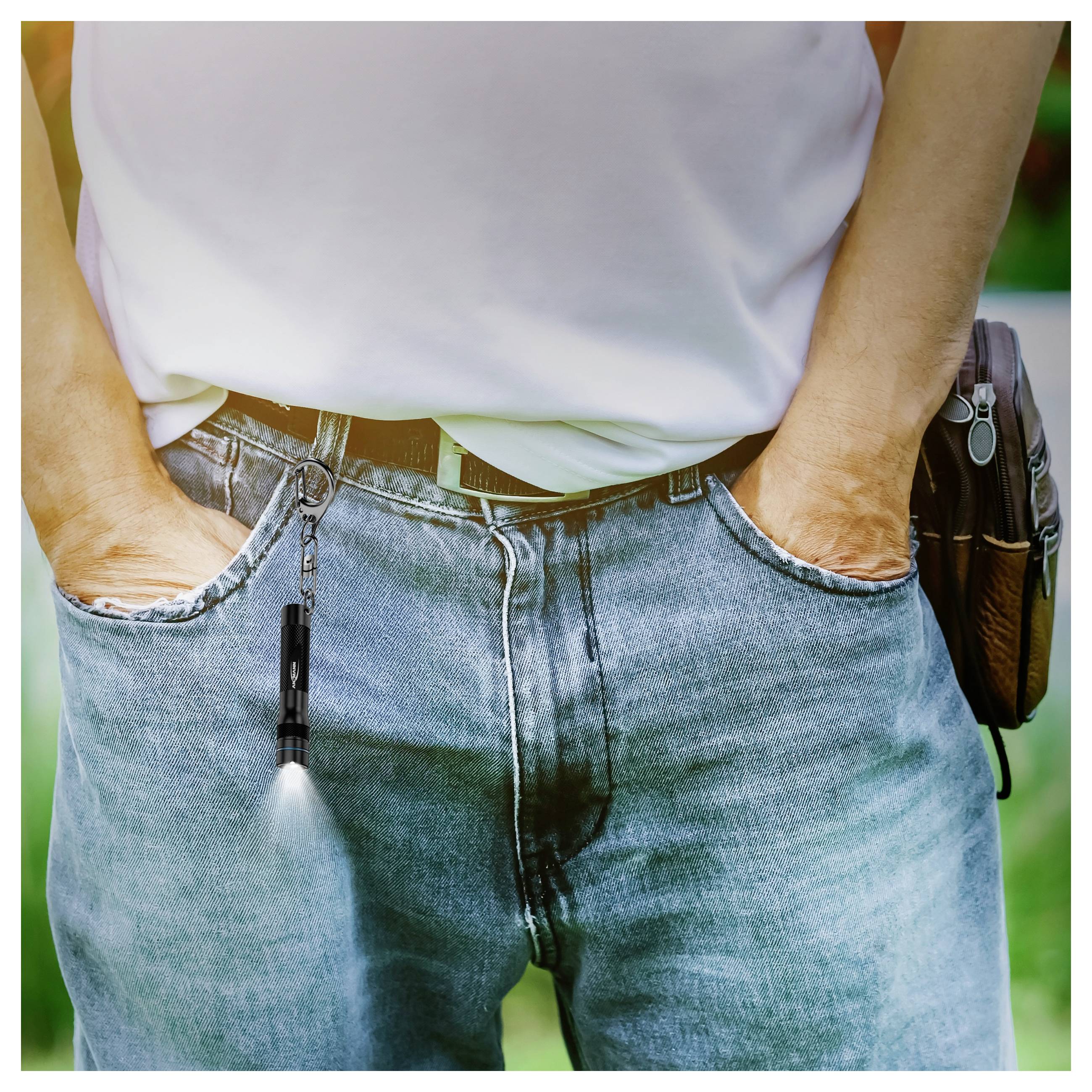 Eine Person in blauen Jeans und einem weißen Hemd steht mit den Händen in den Taschen, wobei eine kleine Taschenlampe an einer Gürtelschlaufe nach unten leuchtet.