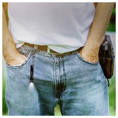 Eine Person in blauen Jeans und einem weißen Hemd steht mit den Händen in den Taschen, wobei eine kleine Taschenlampe an einer Gürtelschlaufe nach unten leuchtet.