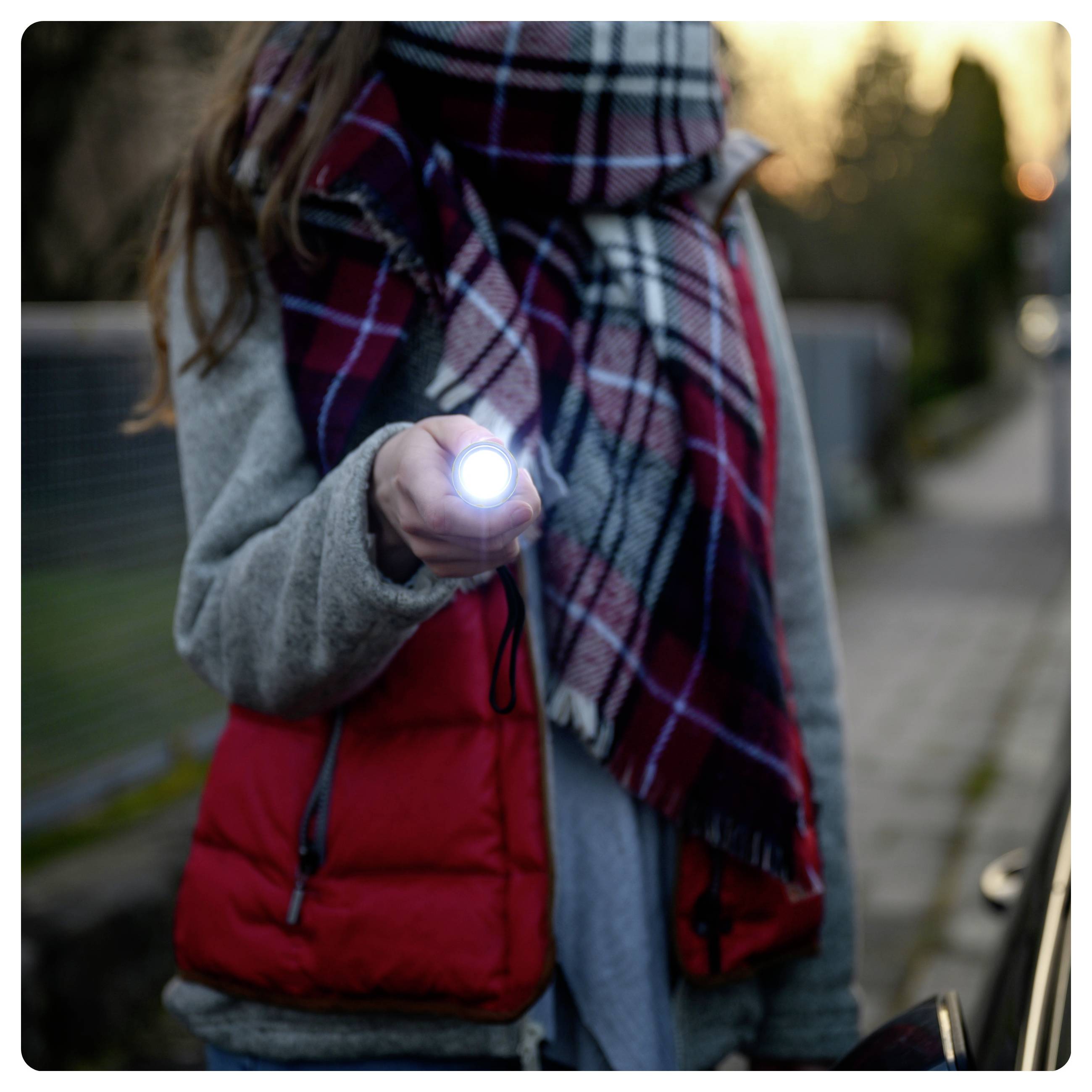Eine Person mit einer roten Weste und einem karierten Schal hält eine eingeschaltete Taschenlampe auf einer nur schwach beleuchteten Straße, was einen Fokus auf Sicherheit oder Erkundung andeutet.