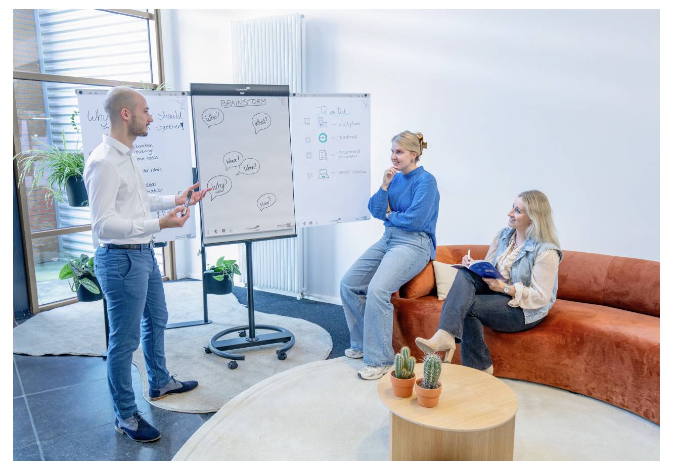 Eine Person steht an einer Whiteboard und zeigt zwei sitzenden Personen ein Konzeptdiagramm in einem modernen, lässigen Büroumfeld.