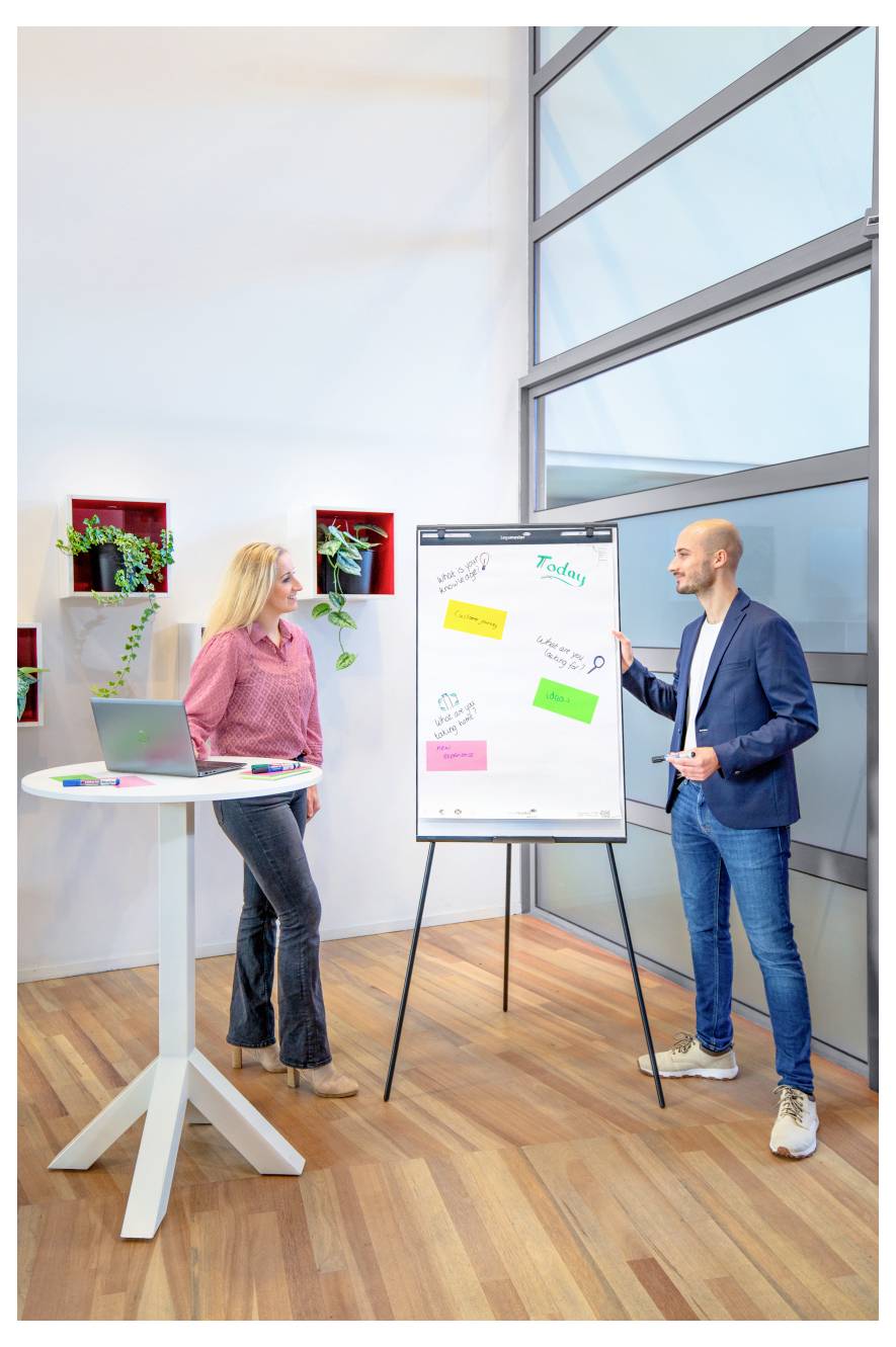 Ein Mann und eine Frau diskutieren in einem hellen Büro Notizen an einem Whiteboard. Die Frau steht neben einem Tisch mit einem Laptop; der Mann gestikuliert auf das Chart.