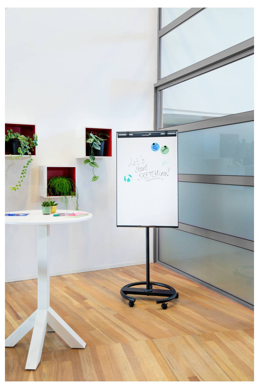 Ein heller Raum mit einem Flipchart, auf dem "Lass uns anfangen zu skizzieren!" geschrieben steht. Auf einem Tisch liegen Marker und eine Pflanze; an den Wandregalen sind weitere Pflanzen ausgestellt.