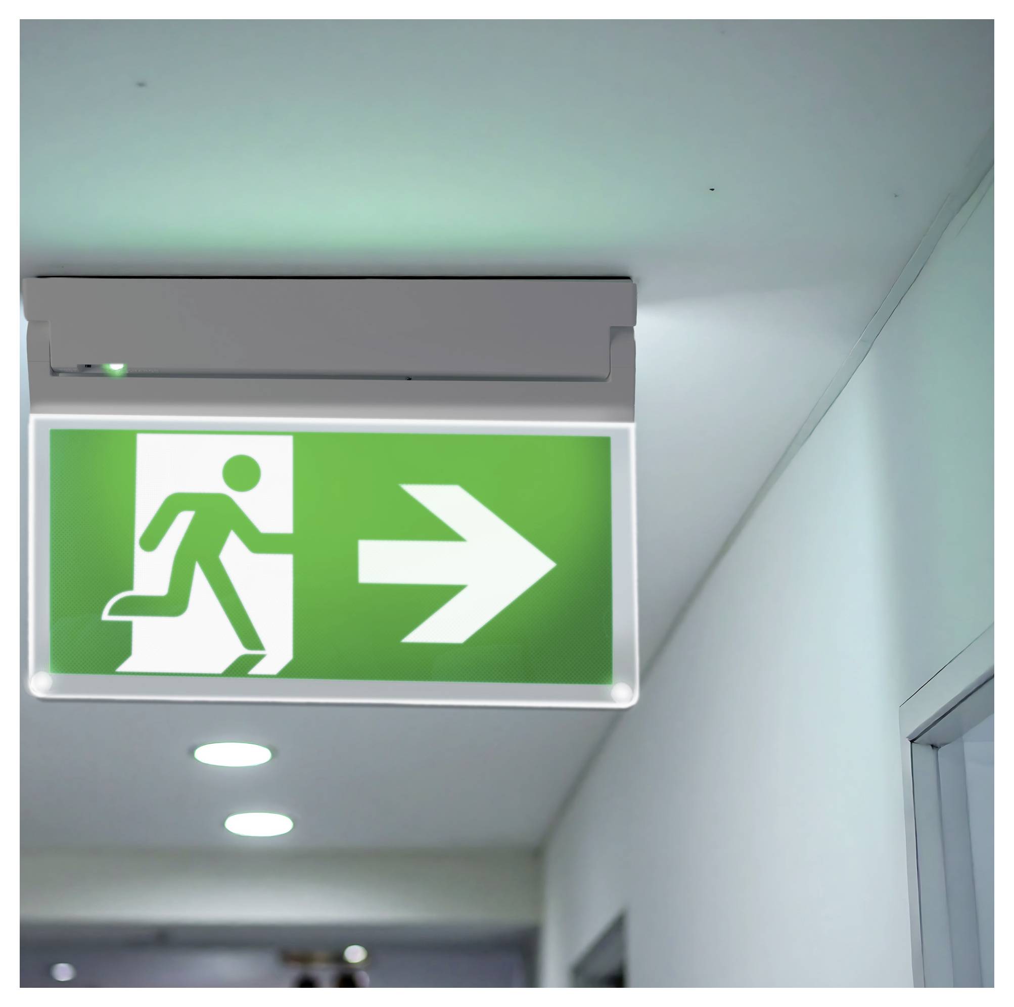 „Grünes Notausgangsschild mit einer Lauf-Figur und einem nach rechts zeigenden Pfeil, an der Decke eines Flurs angebracht. Gibt die Richtung zum Notausgang an."