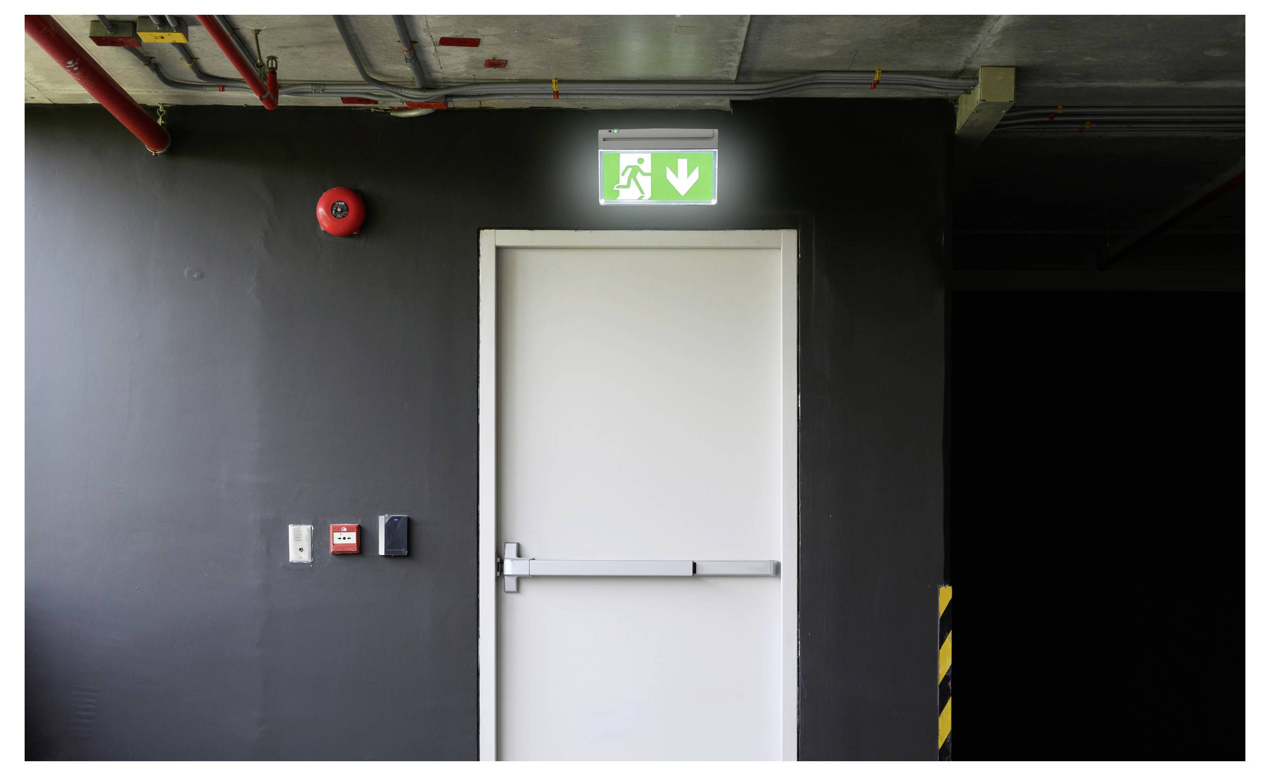 Eine Notausgangstür in einem dunklen Flur mit einem grün beleuchteten "Ausgang"-Schild oben, das eine laufende Figur und einen Pfeil nach unten zeigt.