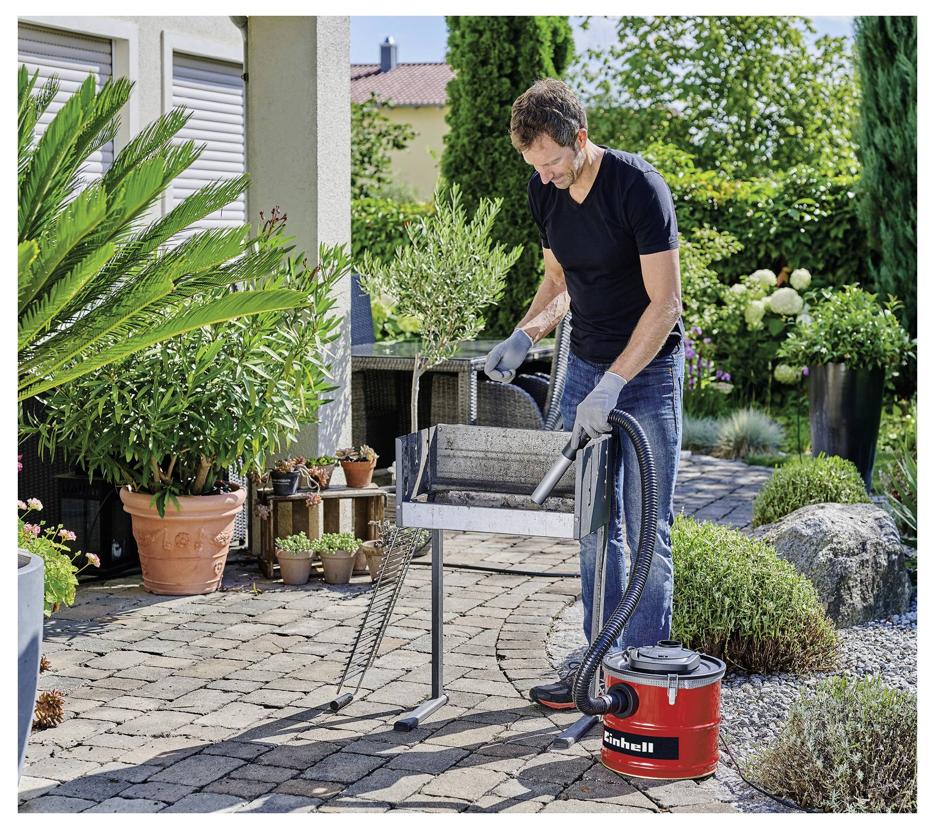 Eine Person reinigt einen Grill mit einem Staubsauger im Freien auf einer Terrasse, umgeben von Pflanzen und Blumentöpfen.