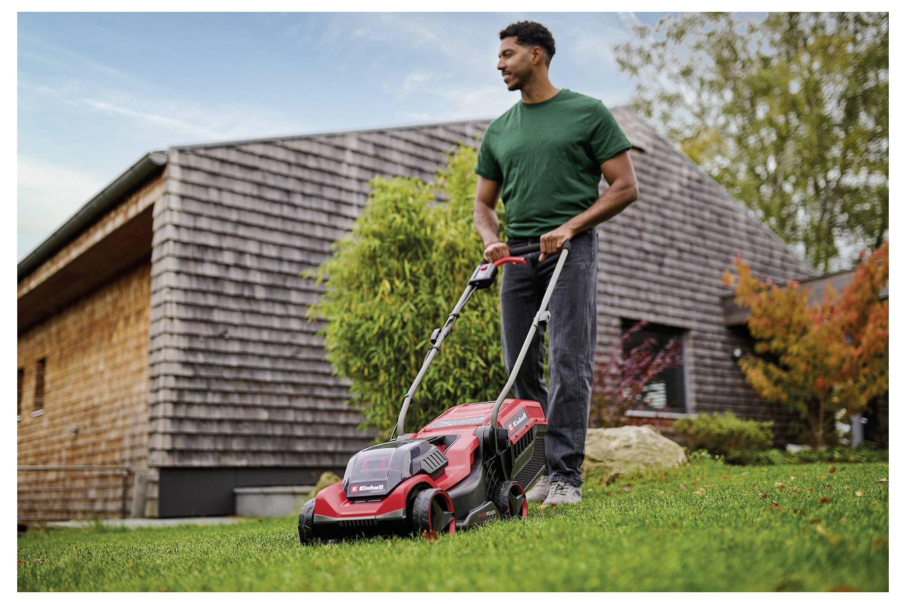 Mann mäht Rasen mit rotem elektrischem Rasenmäher in einem Hinterhof, trägt grünes Hemd und graue Hose, mit einem Holzhaus und Bäumen im Hintergrund.