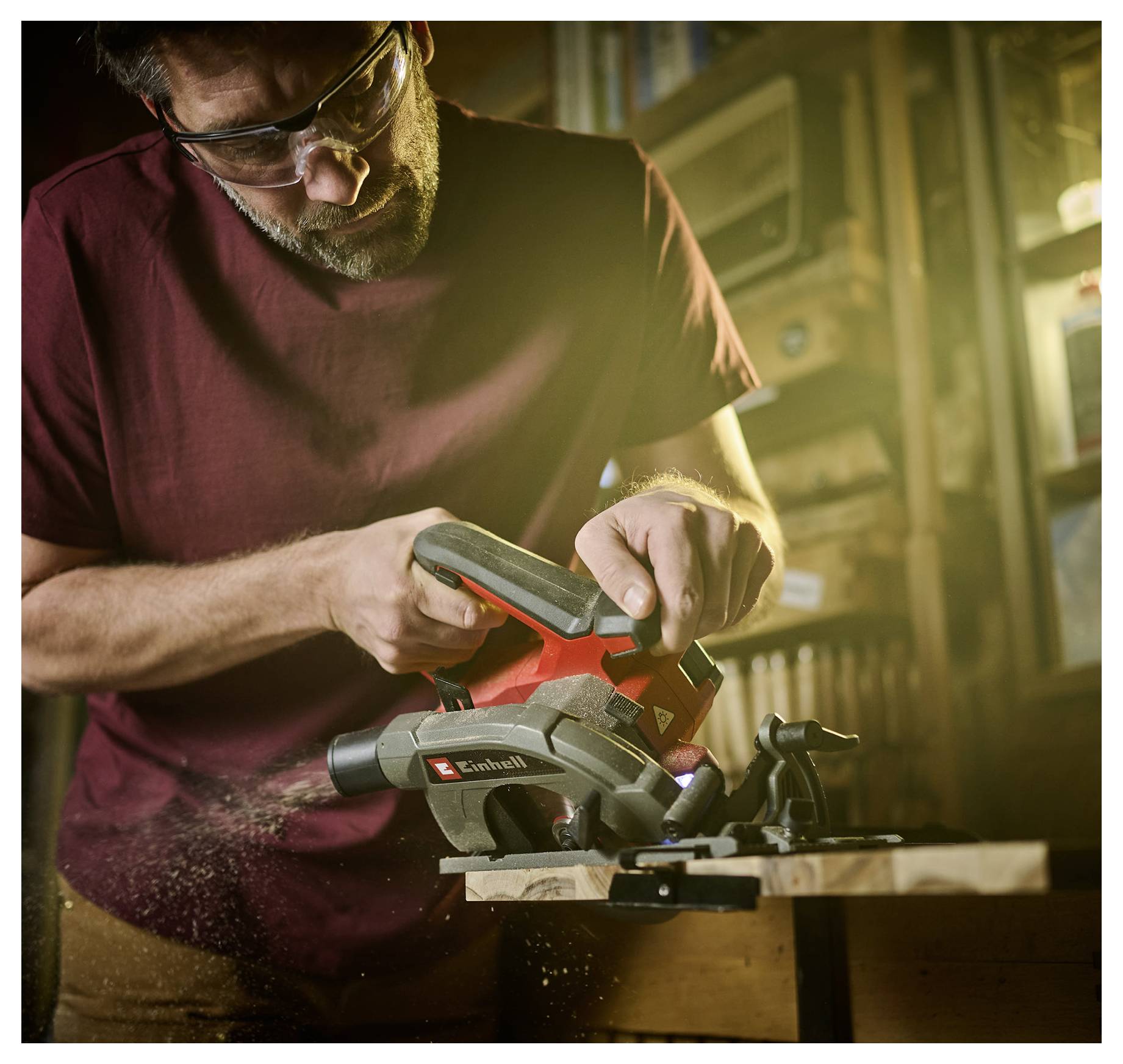 Ein Mann mit Schutzbrille benutzt einen elektrischen Handhobel an einem Holzbrett in einer Werkstatt, während Holzspäne umherfliegen.