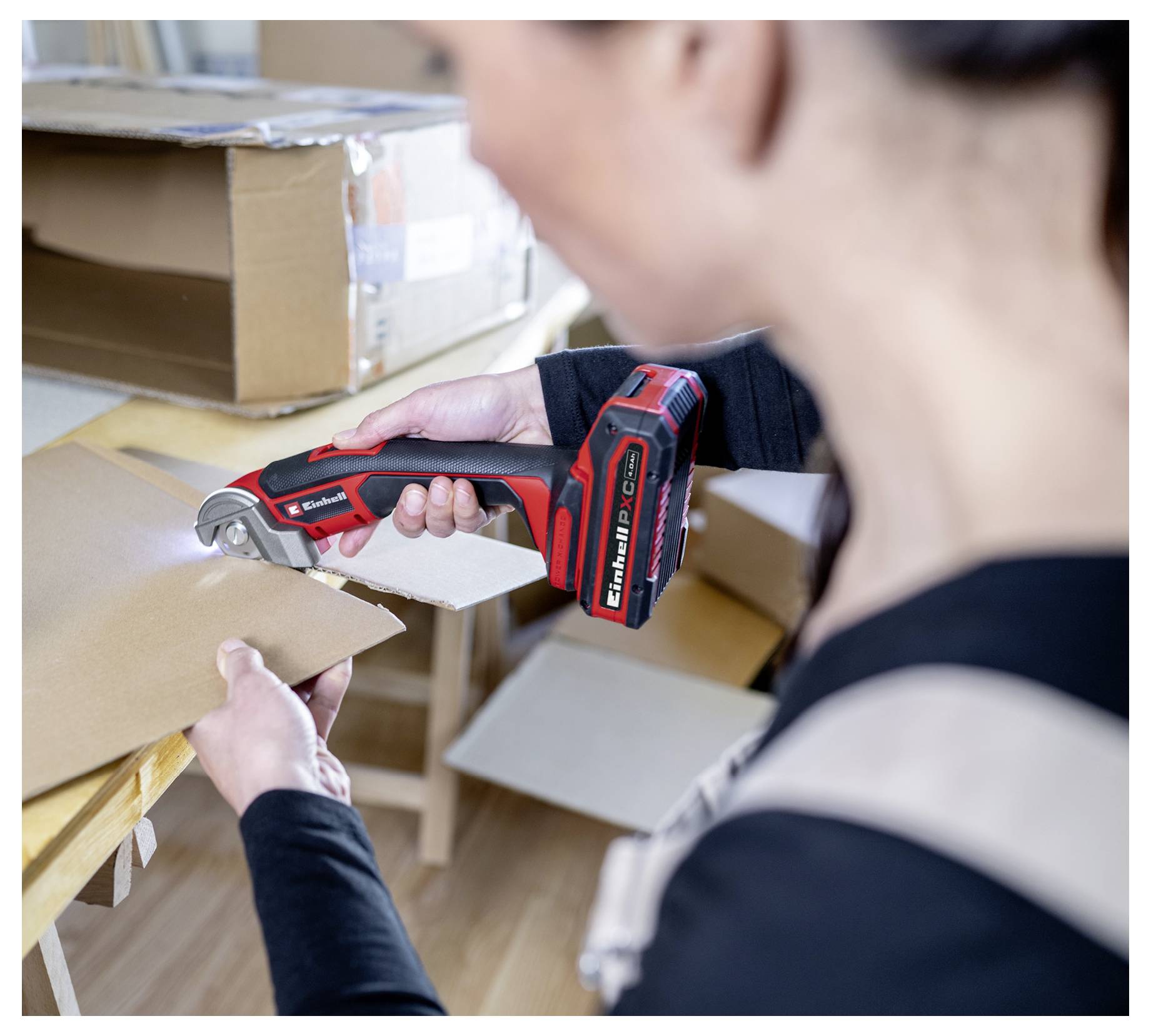 Eine Person benutzt ein rot-schwarzes elektrisches Werkzeug, um Karton zu schneiden. Im Hintergrund sind Kartonschachteln zu sehen.