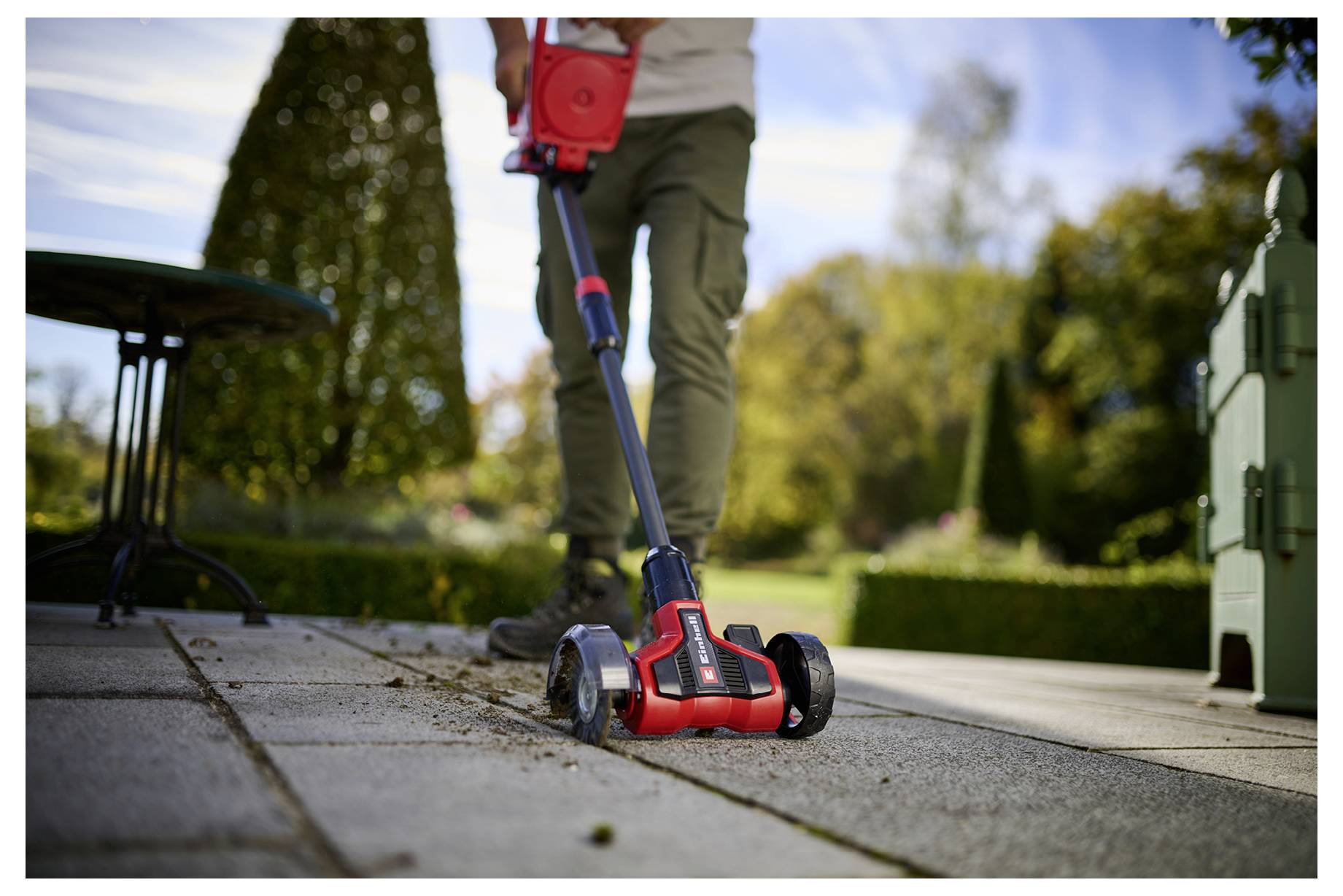 Eine Person benutzt einen roten und schwarzen elektrischen Besen auf einer Terrasse und beseitigt dabei Schmutz. Im unscharfen Hintergrund sind Bäume und Gartenmöbel zu erkennen.