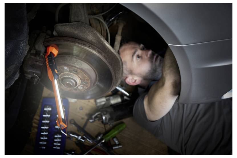 Ein Mechaniker liegt unter einem Auto und untersucht die Bremsen. Eine orangefarbene Arbeitslampe beleuchtet den Bereich, während Werkzeuge am Boden verstreut liegen.