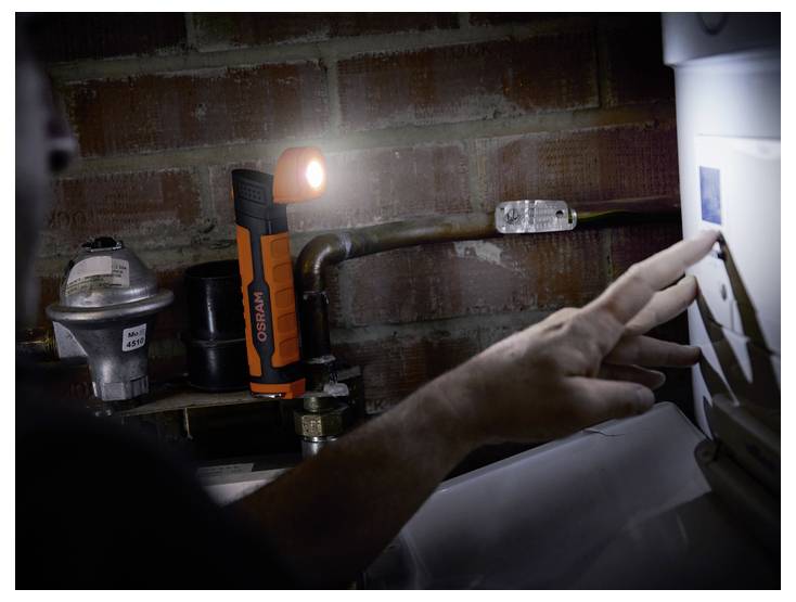 Eine Person justiert die Einstellungen an einem Wassererhitzer in einem nur schwach beleuchteten Raum und verwendet dabei eine orangefarbene LED-Taschenlampe für bessere Sichtbarkeit.