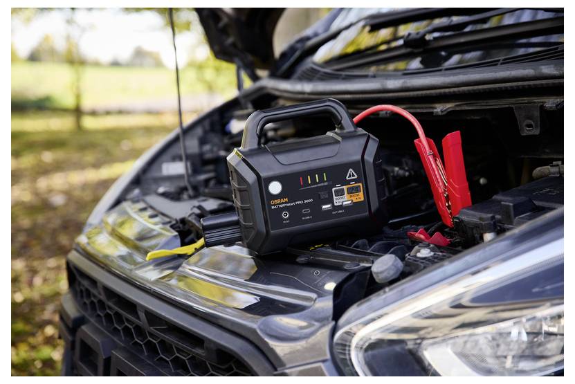 Ein tragbarer Autobatterieanlasser mit angeschlossenen roten und schwarzen Kabeln ist in einer Außenumgebung mit einem Automobildmotor verbunden.