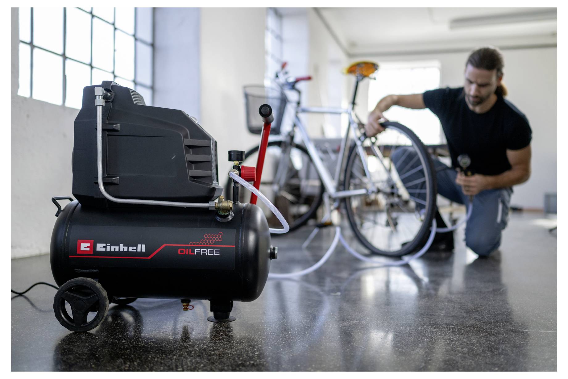Ein Mann kniet in einer Garage und repariert ein Fahrrad neben einem schwarzen ölfreien Einhell-Kompressor im Vordergrund.