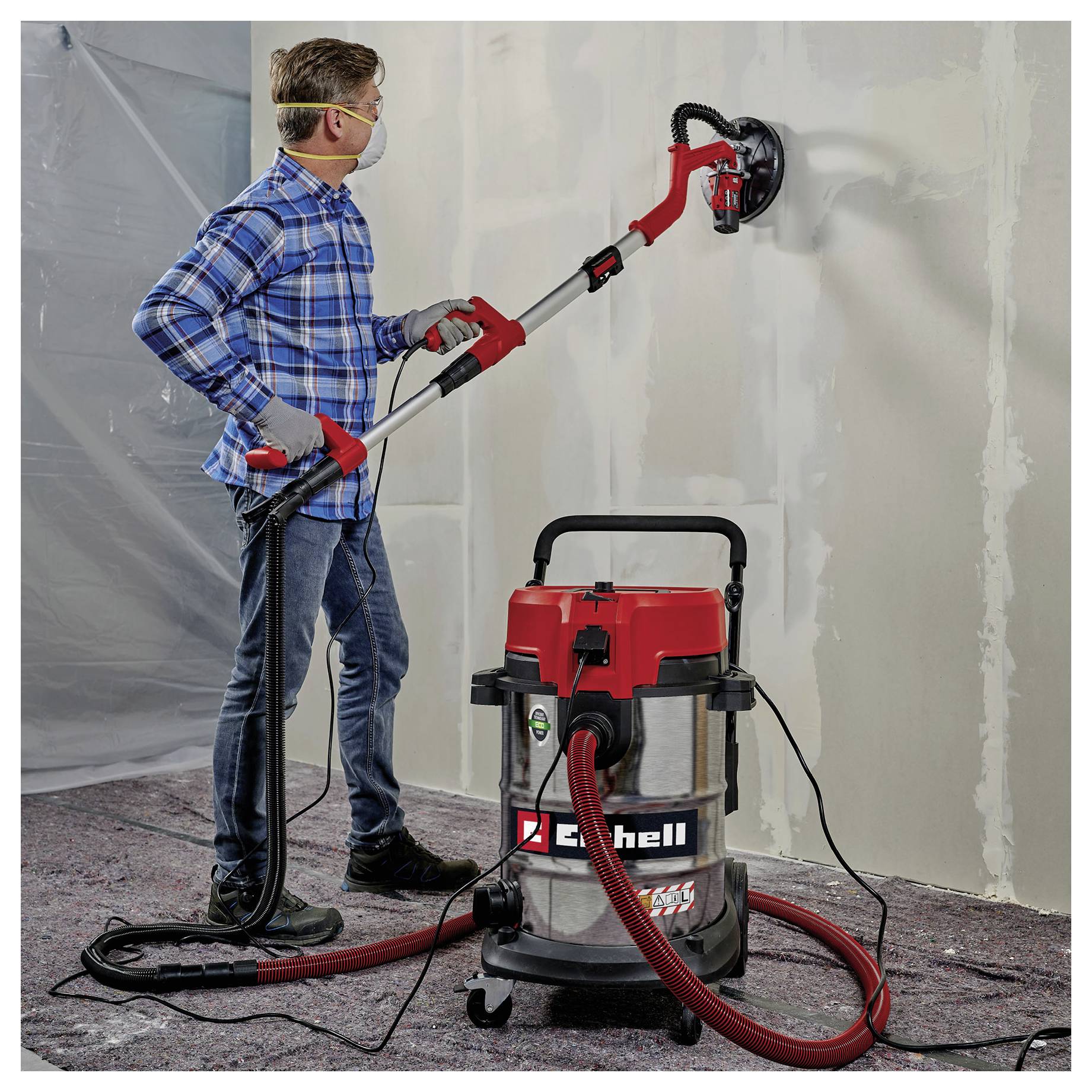 Eine Person, die eine Wand mit einem elektrischen Trockenbauschleifer, der an einen Staubsauger angeschlossen ist, schleift und dabei Schutzausrüstung in einer Renovierungsumgebung trägt.