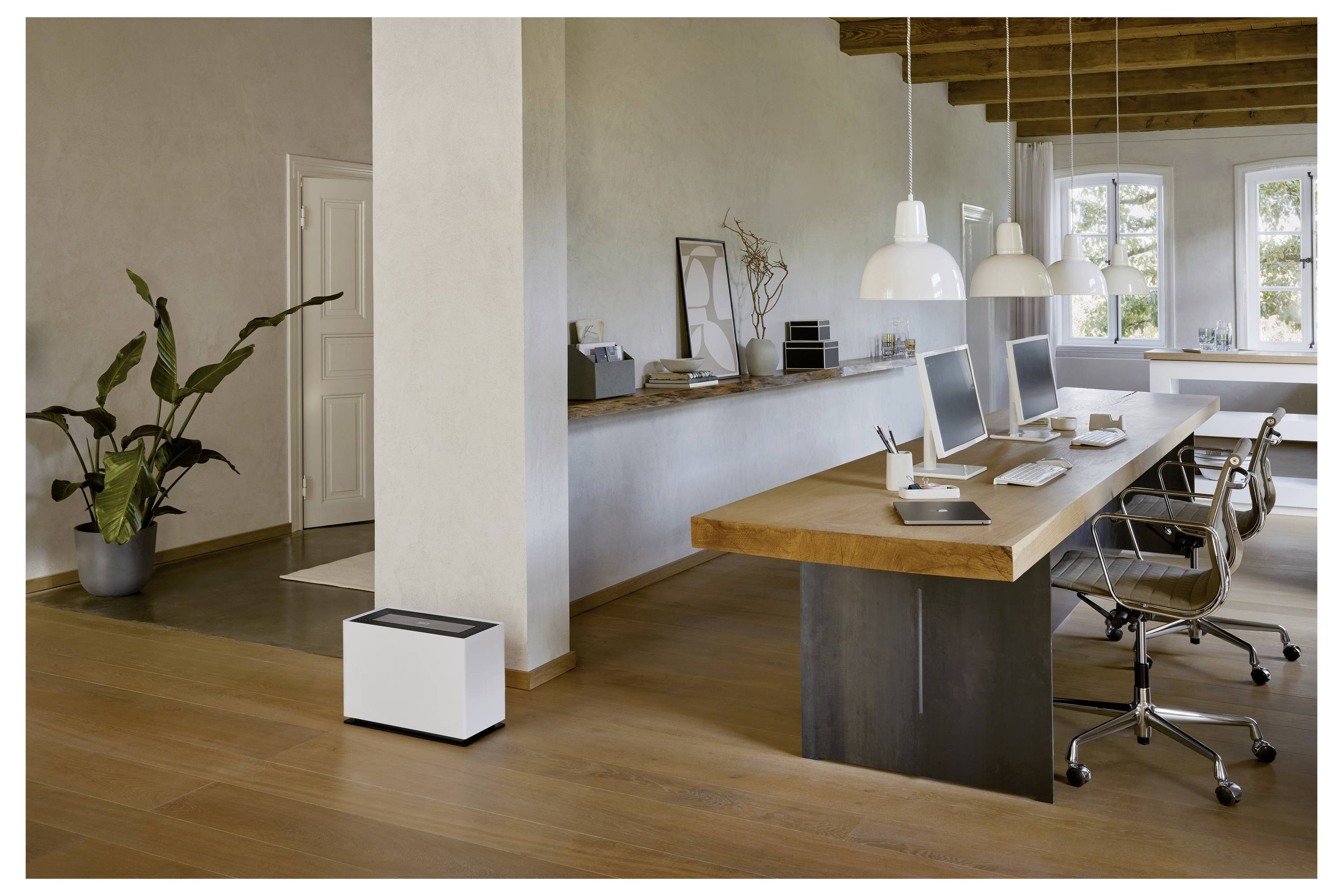 Ein modernes Büro mit einem großen Holzschreibtisch, vier Stühlen, drei Computern und hängenden Lampen. Eine Pflanze und ein weißes Gerät befinden sich auf dem Boden.