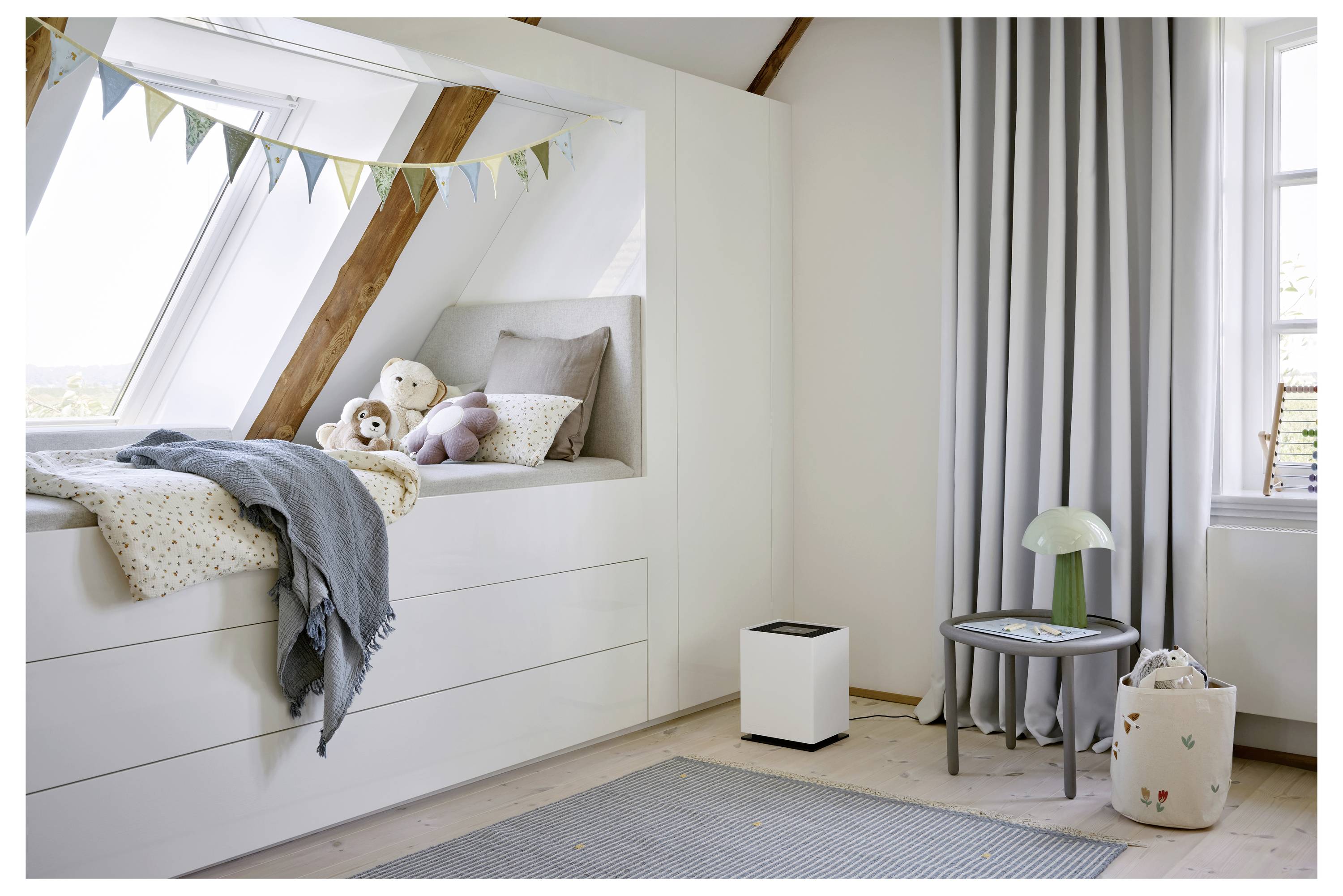 Ein gemütliches Kinderzimmer mit einem Tagesbett unter einem schrägen Fenster, geschmückt mit Wimpelkette. Weiche Kuscheltiere und Kissen ruhen auf dem Bett. Ein kleiner Tisch mit einer Lampe und einem Korb steht in der Nähe.
