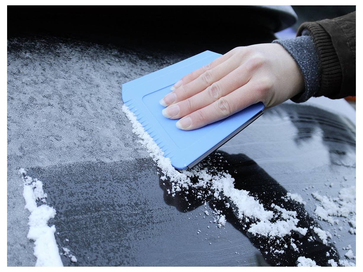 Eine Person verwendet einen blauen Eiskratzer, um Eis von der Windschutzscheibe eines Autos zu entfernen, was auf eine kalte Winterumgebung hindeutet.