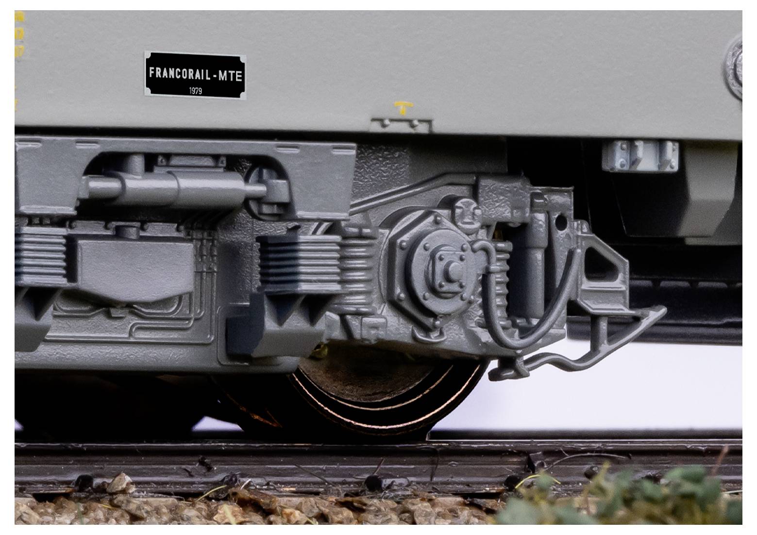 Nahaufnahme eines Modelleisenbahnrades und Federungsdetails auf einer Schiene, mit einem kleinen Schild im Hintergrund, auf dem "Francorail-MTE 1979" steht.