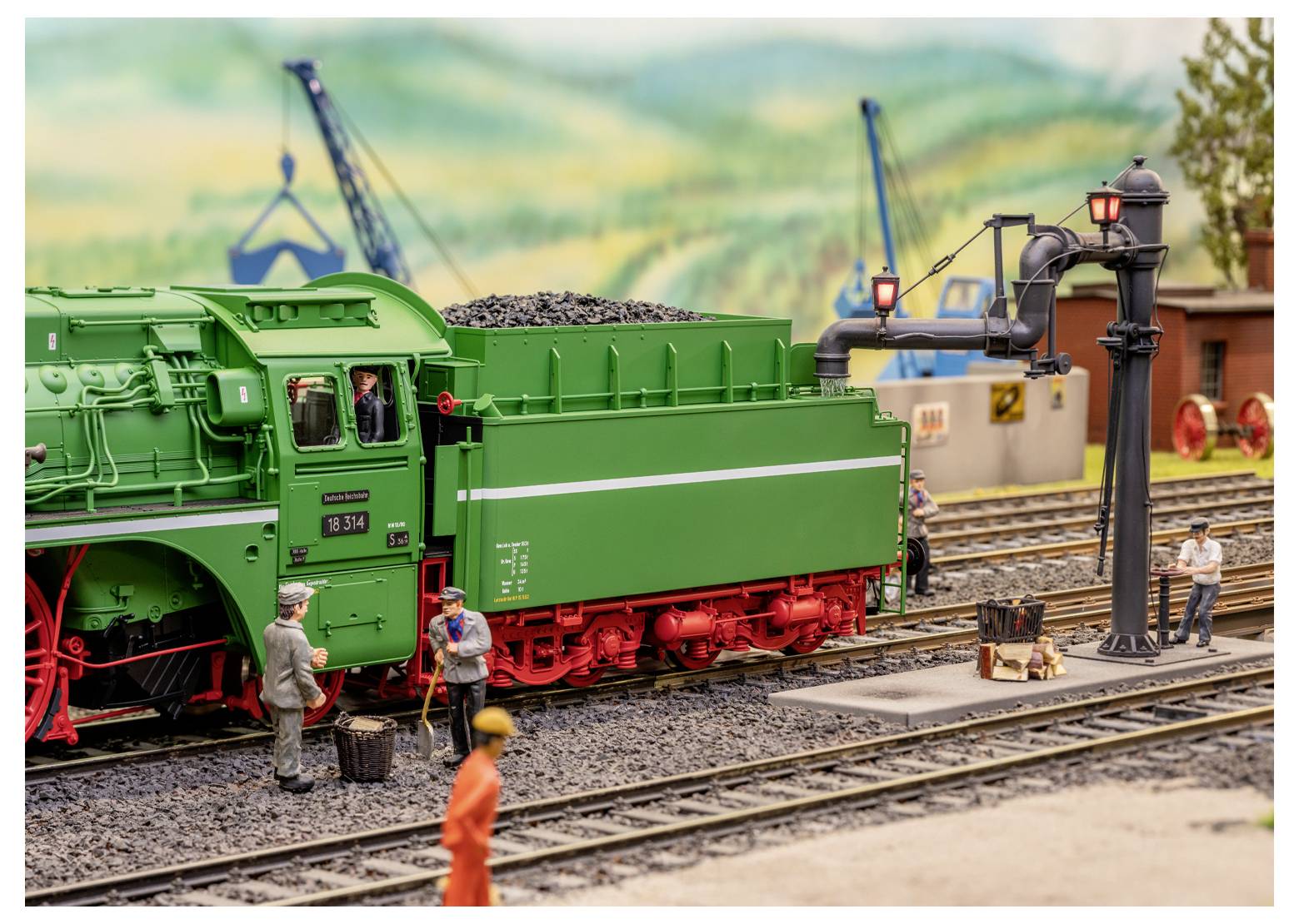 Eine Modellbahn-Diorama zeigt eine grüne Dampflokomotive auf Schienen, mit Arbeitern in der Nähe und Kranen im Hintergrund.