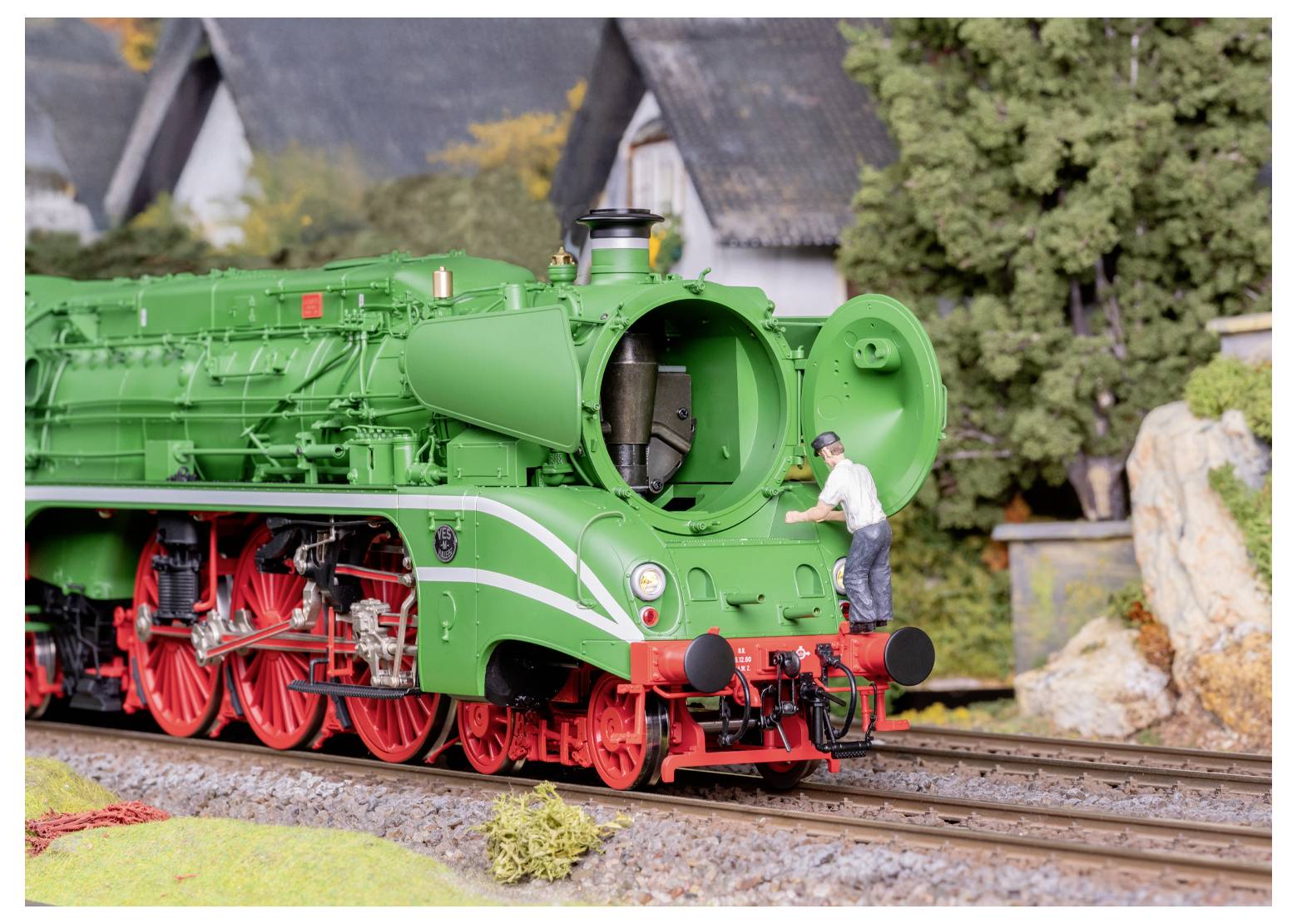 Eine Modelleisenbahnszene zeigt einen Mann, der die Front einer grünen Dampflokomotive inspiziert und dabei die detaillierte Handwerkskunst und Realitätsnähe hervorhebt.
