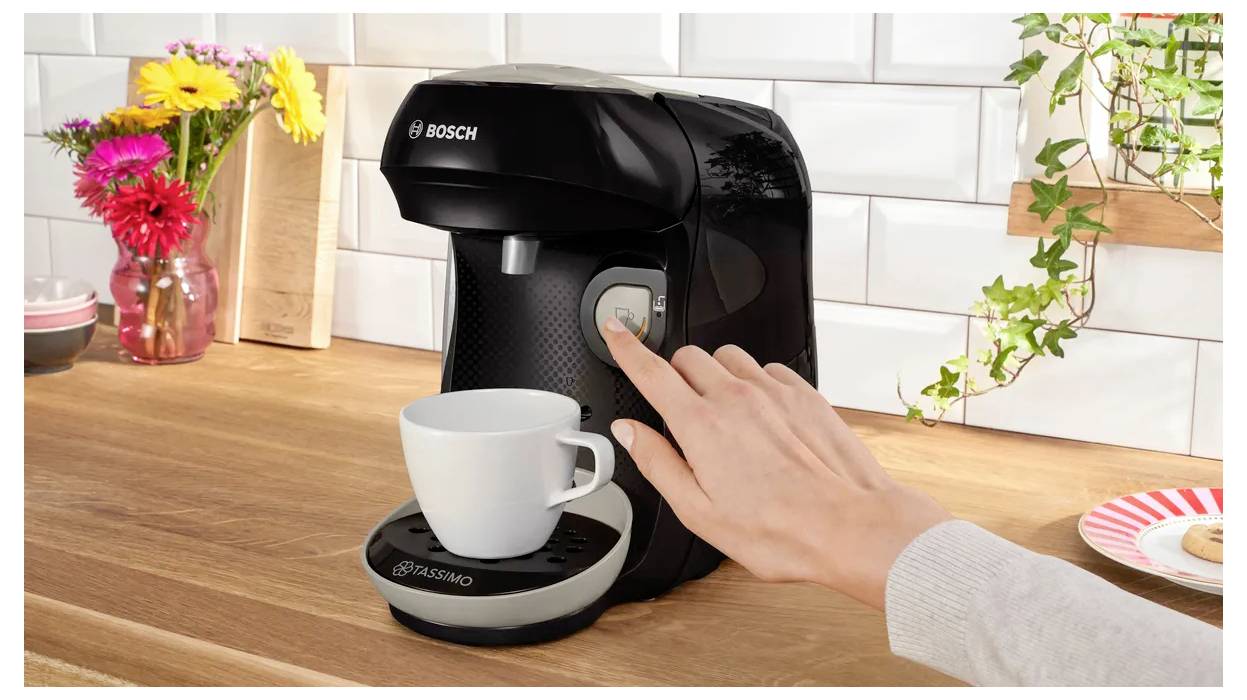 Eine Person drückt einen Knopf an einer schwarzen Kaffeemaschine; eine weiße Tasse wartet darunter am Ausguss. Bunte Blumen und Tassen befinden sich im Hintergrund.