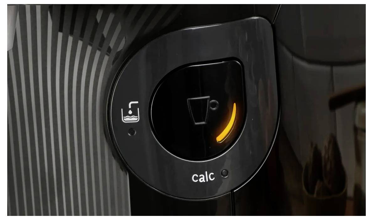 Eine Nahaufnahme des Anzeigefelds einer Kaffeemaschine, die ein leuchtendes orangefarbenes Licht über der Beschriftung "calc" neben einem Wassertanksymbol zeigt.