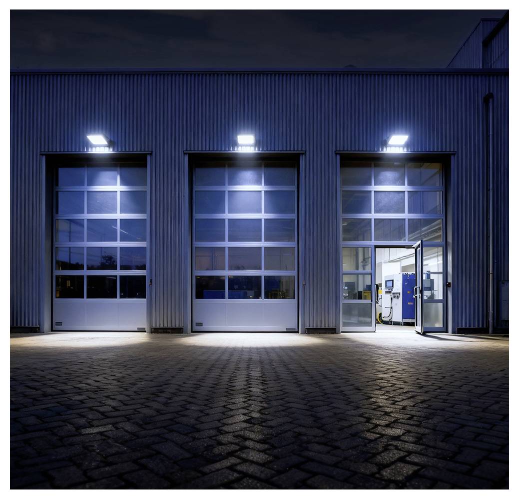 Ein Industriegebäude mit drei großen Garagentoren unter hellen Lichtern bei Nacht, wobei eine Tür geöffnet ist und im Inneren Maschinen sichtbar macht.
