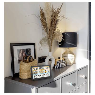 Ein Sideboard mit einer dekorativen Lampe, gerahmtem Foto, Flechtkörbchen und einem digitalen Display, das Uhrzeit, Datum, Wetter und Temperatur anzeigt.