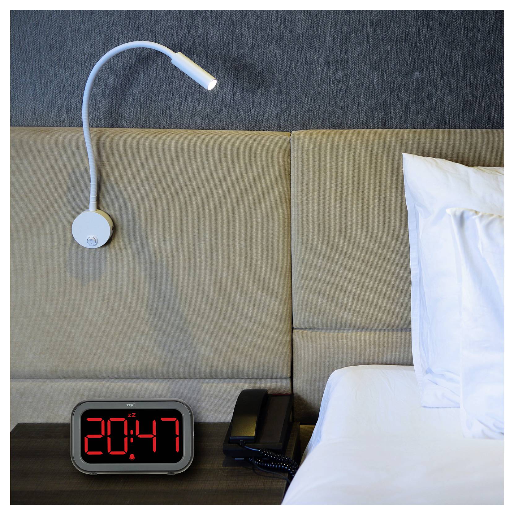 Ein Bett mit weißen Kissen, eine flexible Leselampe an einer gepolsterten Wand und ein Wecker, der '20:47' anzeigt, neben einem Telefon.