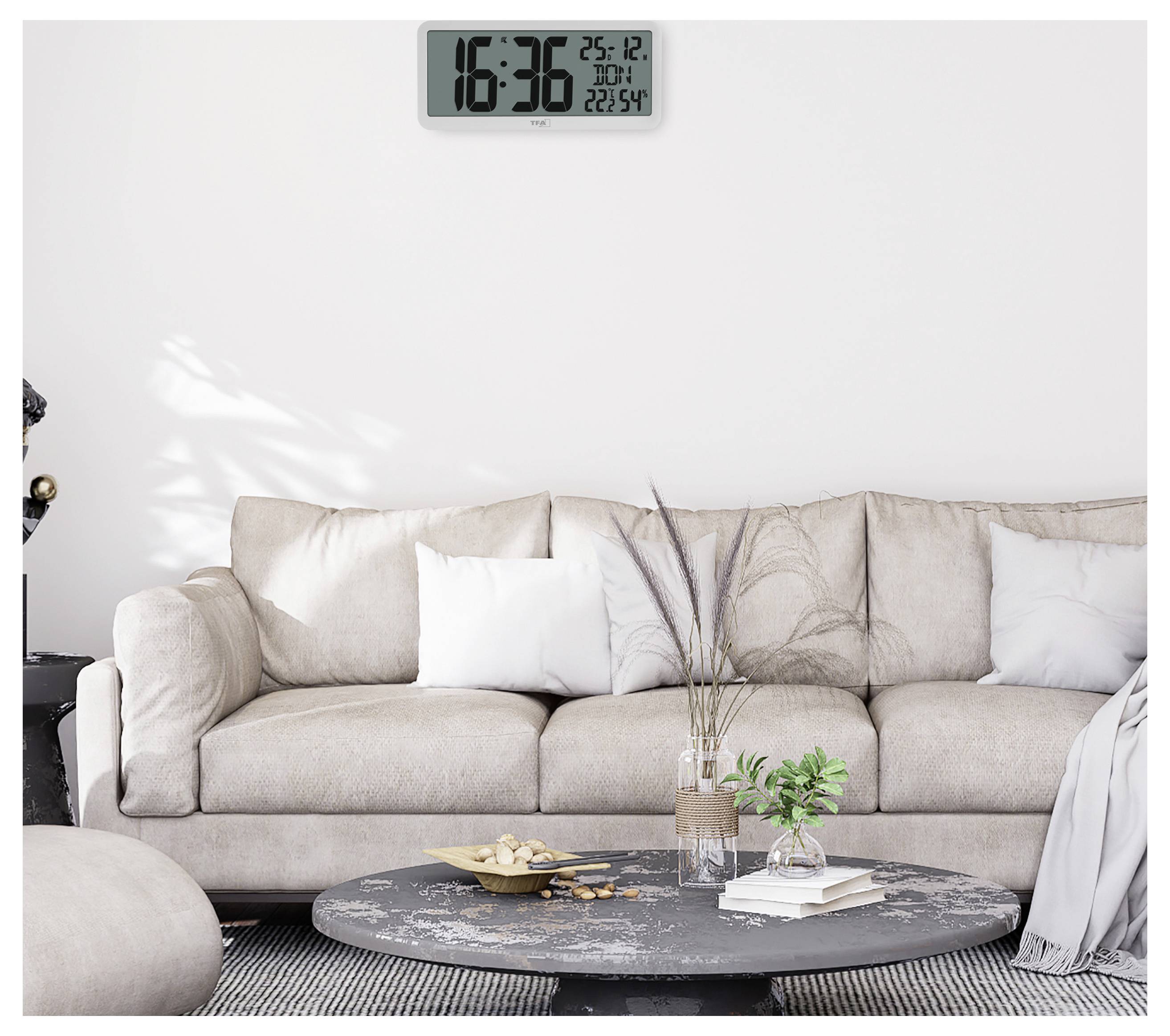 Ein modernes Wohnzimmer zeigt ein beiges Sofa, einen runden schwarzen Couchtisch mit Pflanzen und eine digitale Wanduhr, die "16:36, 25-12" anzeigt.