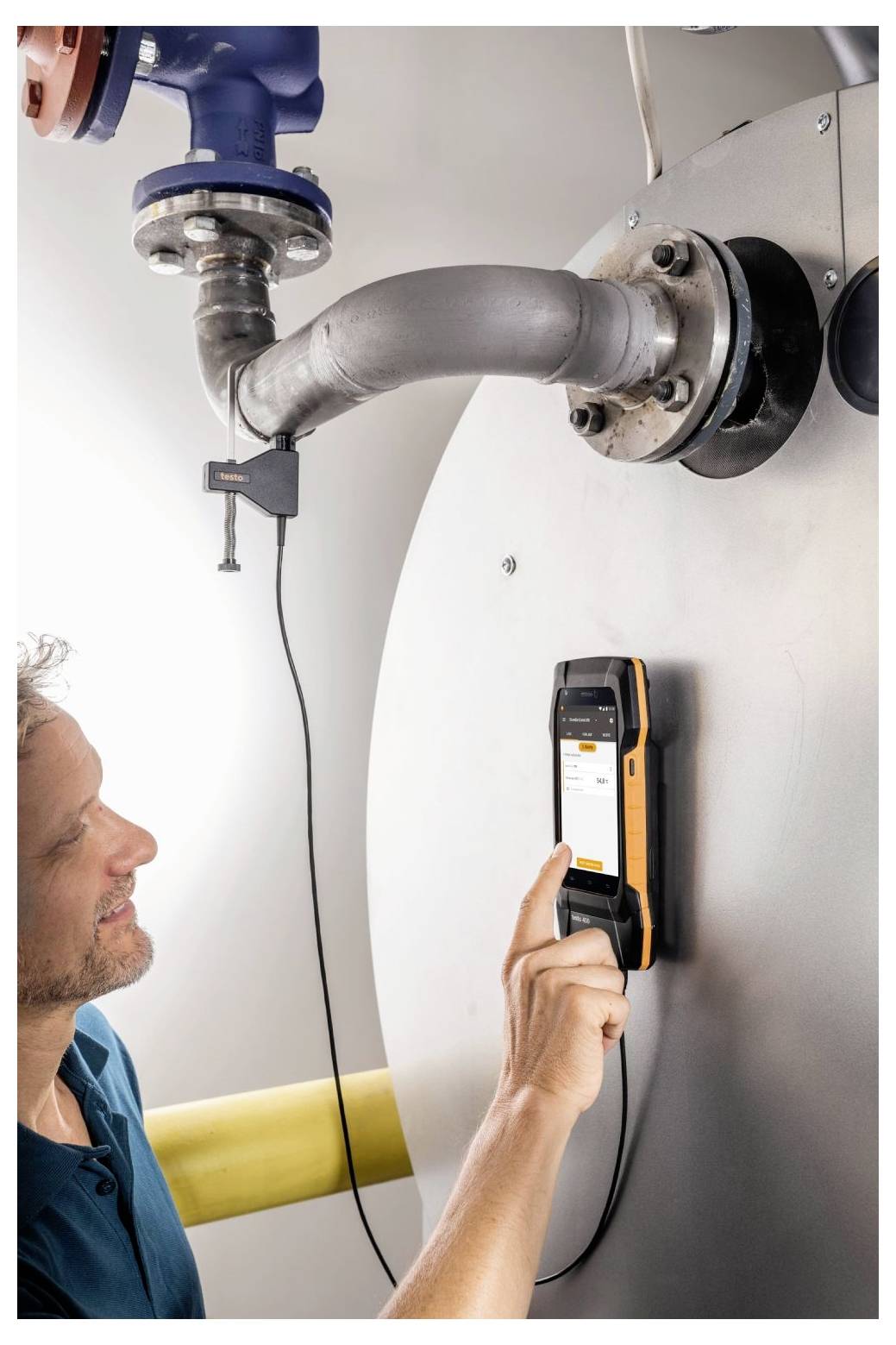 Person mit einem Handgerät zur Messung von Emissionen aus einem Metallrohr. Das Gerät ist am Rohr angebracht und zeigt Daten an.
