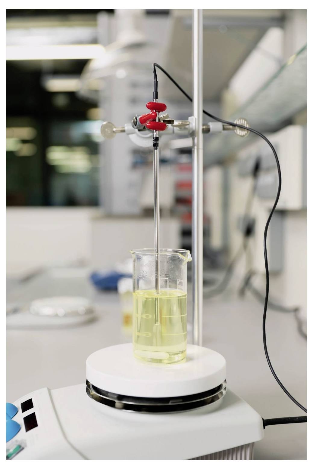 Ein Becherglas mit gelber Flüssigkeit auf einem Magnetrührer mit einem Thermometer und einer Stativklemme in einer Laborumgebung.