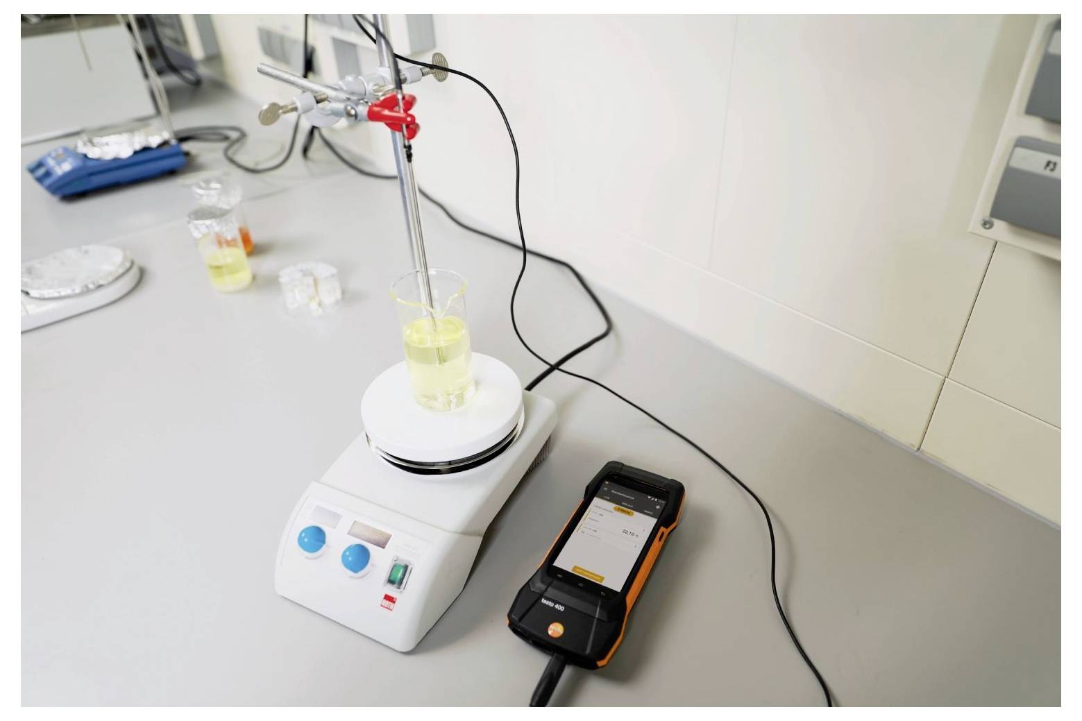 Ein Laboraufbau zeigt einen Becher auf einem Magnetrührer mit einem digitalen Thermometer, das die Temperatur der Flüssigkeit misst. Ausrüstung umgibt ihn.