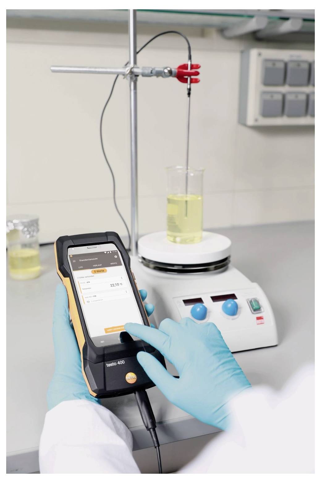 Eine Person in einem Labor trägt Handschuhe und verwendet einen digitalen Messgerät, um den pH-Wert einer gelben Flüssigkeit in einem Becherglas auf einem Magnetrührer zu messen.