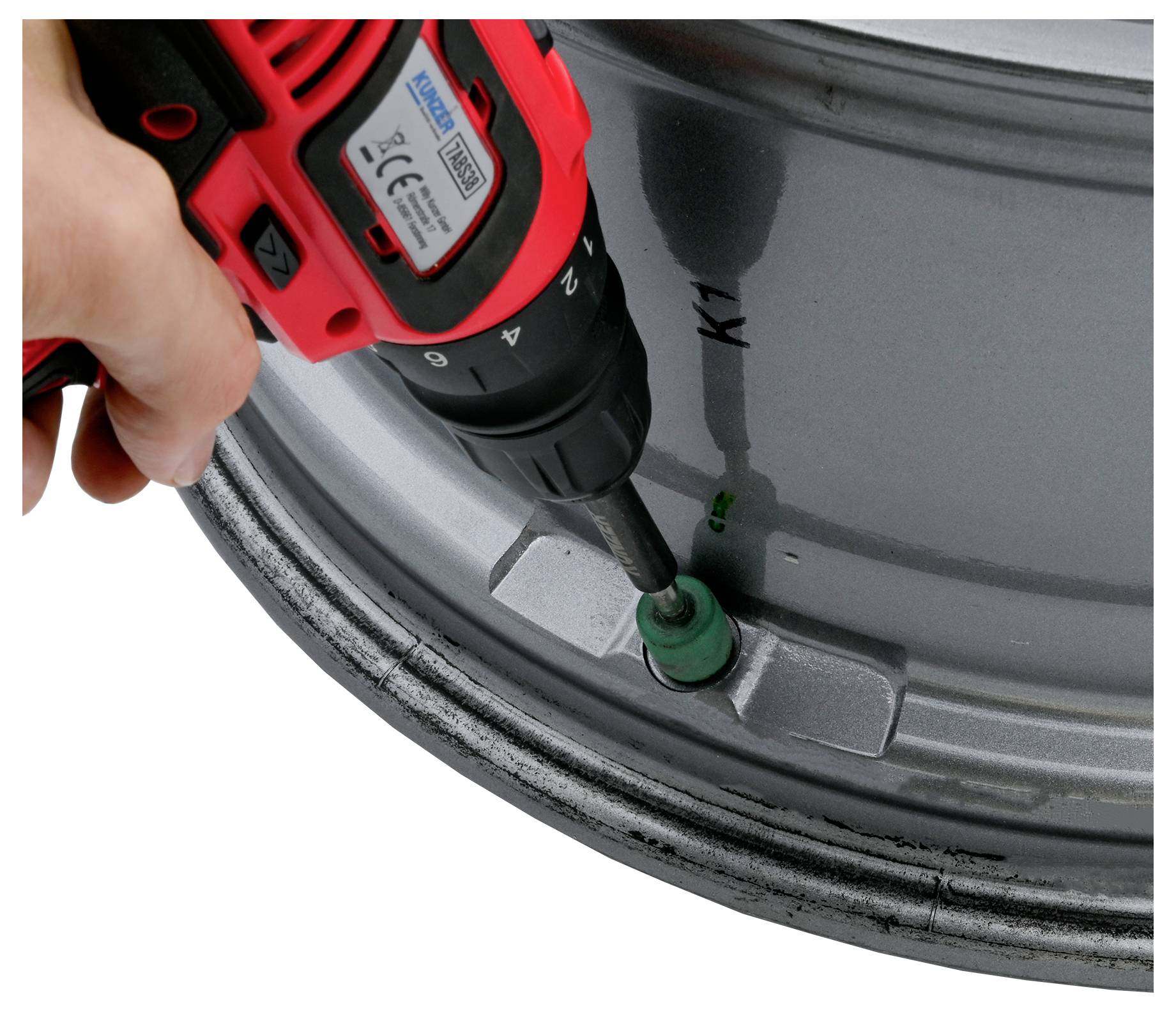 Eine Hand, die einen roten Akkuschrauber verwendet, um ein grünes Ventil an einem großen metallischen Objekt zu reparieren oder zu befestigen, möglicherweise eine Felge oder ein Rad, markiert mit K1.