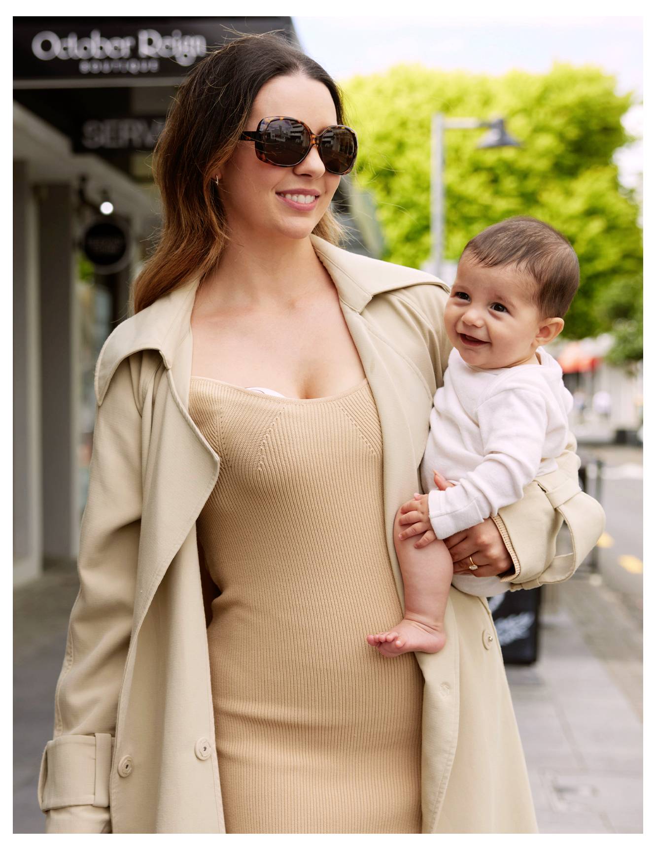 Eine Frau mit Sonnenbrille und Trenchcoat geht im Freien, ein lächelndes Baby auf dem Arm. Helles Tageslicht; Bäume und Geschäfte im Hintergrund.