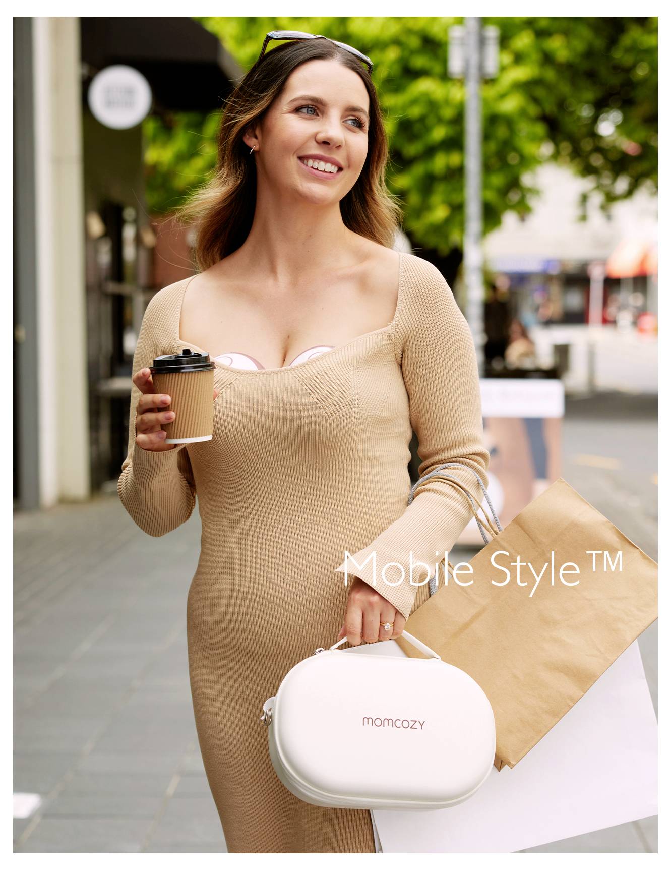 Eine lächelnde Frau im beigen Kleid trägt Einkaufstüten und eine Kaffeetasse und geht im Freien spazieren. Sie hat eine weiße 'Momcozy' Tasche bei sich.