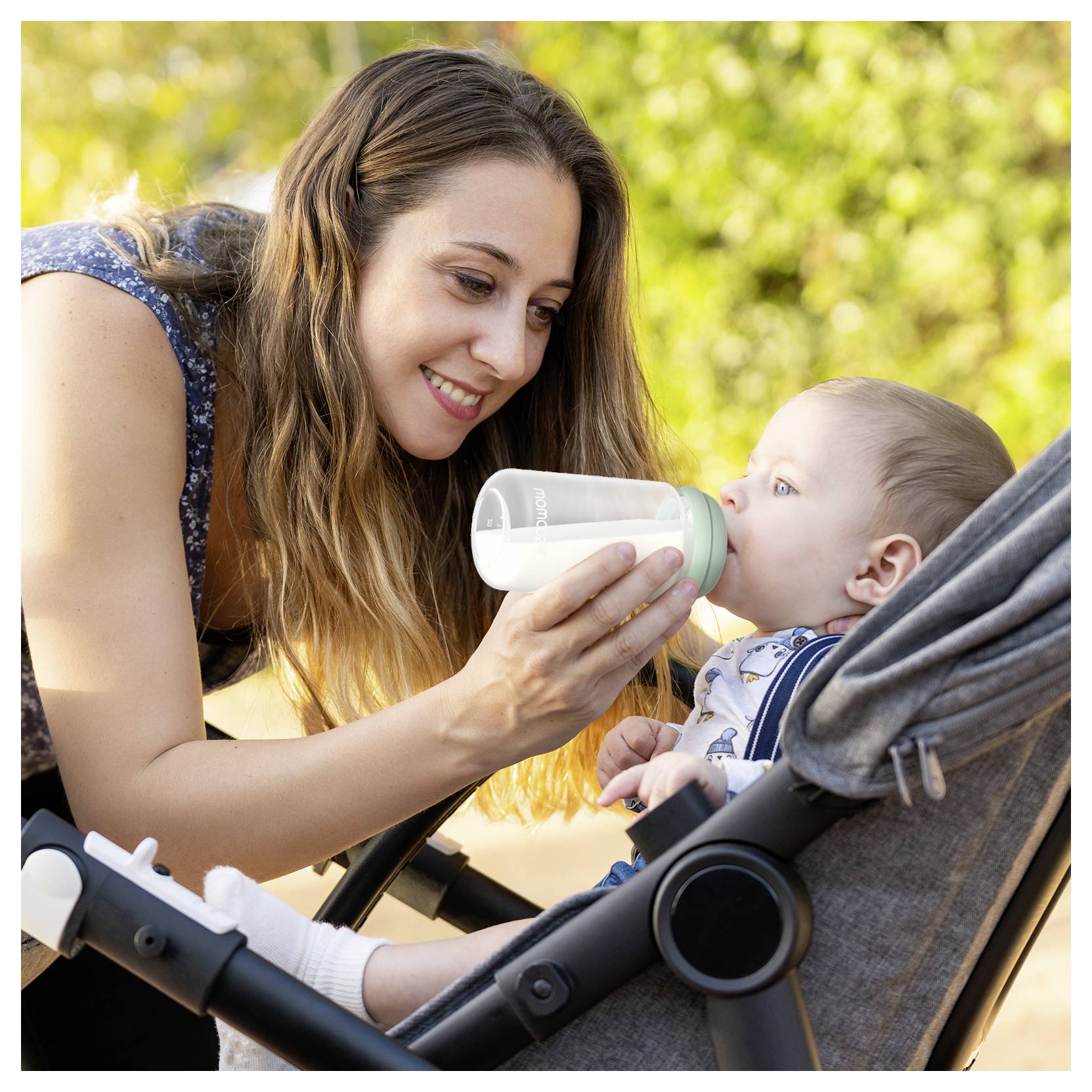 Eine Frau füttert sanft ein Baby mit einer Flasche, während das Baby in einem Kinderwagen sitzt. Sie befinden sich im Freien, umgeben von Grünpflanzen.