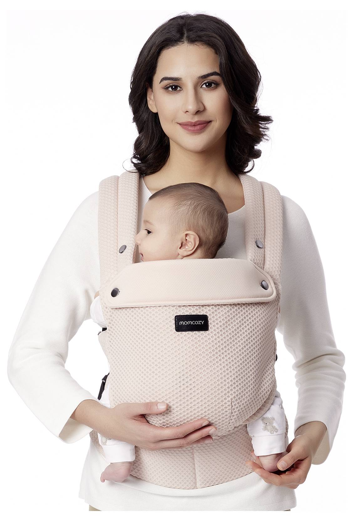 Eine Frau trägt einen beigen Babytrage und hält ein Baby in Vorwärtsposition. Die Frau lächelt und trägt ein weißes Oberteil.