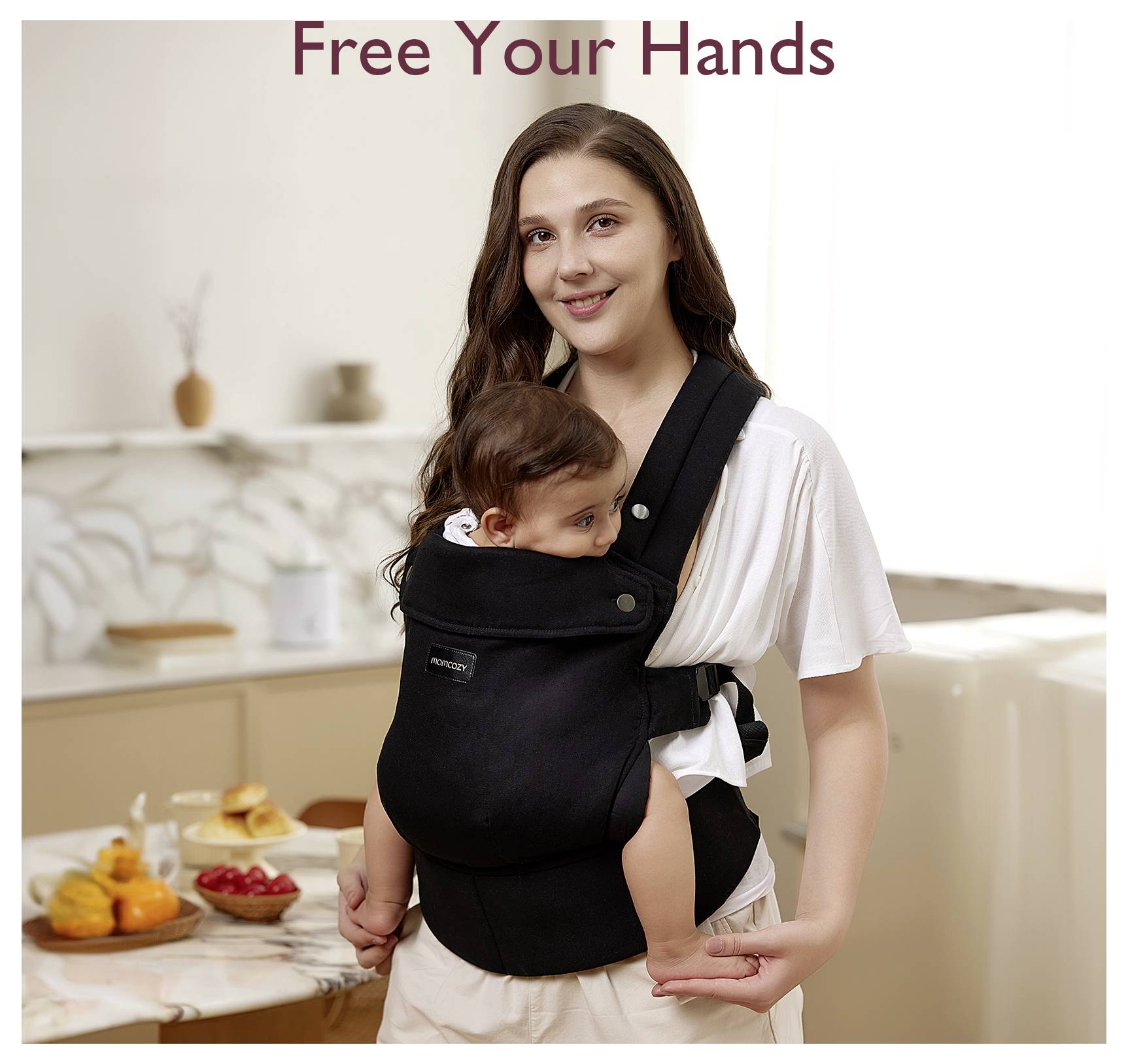 Eine Frau lächelt, während sie ein Baby in einem schwarzen Babytrage in einer Küche trägt. Der Text darüber sagt „Befreie Deine Hände".