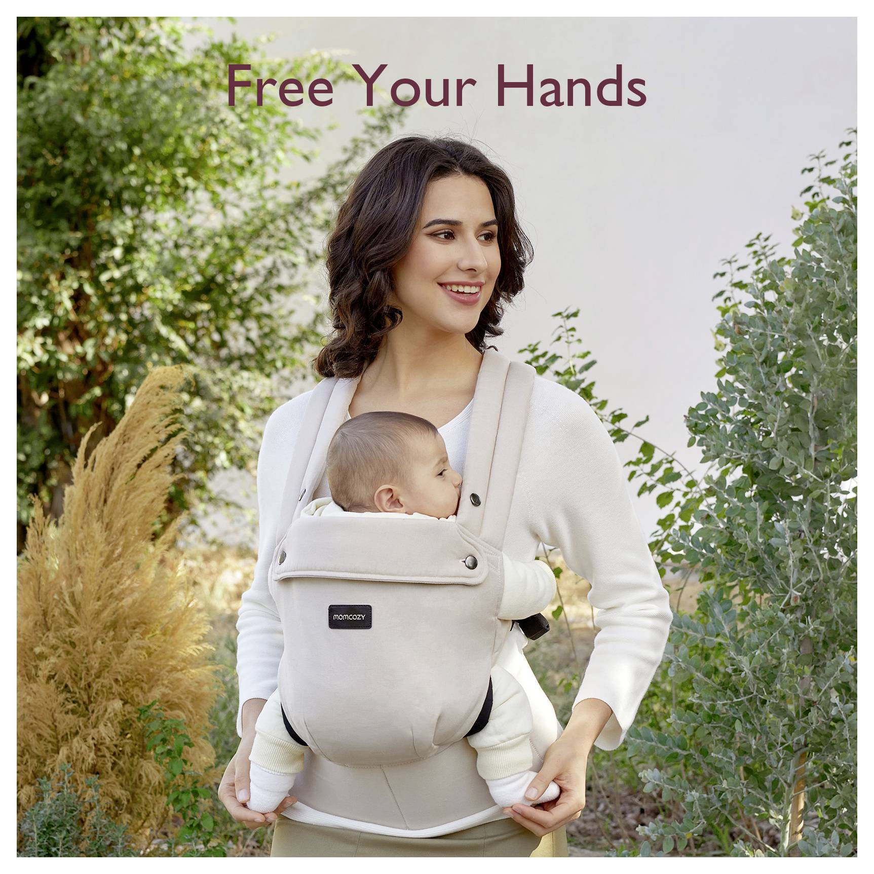 Eine Frau steht im Freien und lächelt, während sie ein Baby in einem beigen, nach vorne gerichteten Babytrage trägt. Der Text darüber liest: "Befreie deine Hände".