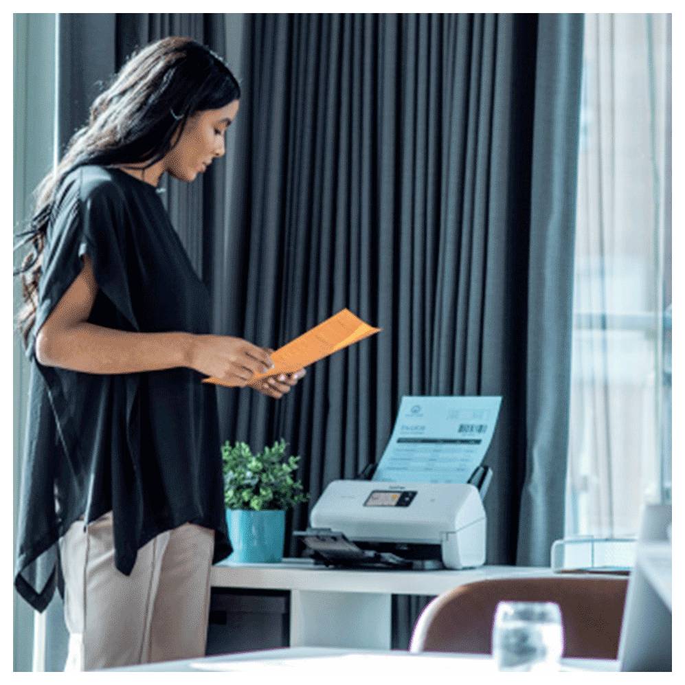 Eine Frau steht in einem Büro und hält ein Dokument vor einem Drucker auf einem Schreibtisch, während im Hintergrund große Fenster und eine Pflanze zu sehen sind.