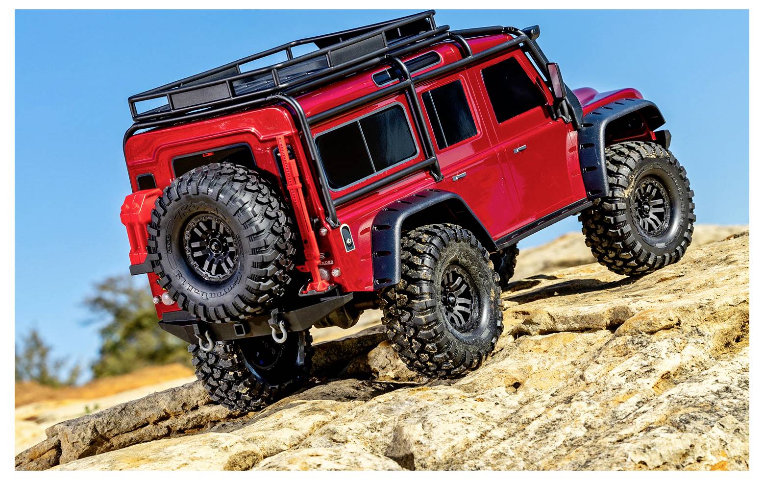 Ein roter Geländespielzeugwagen erklimmt felsiges Gelände unter einem klaren blauen Himmel. Robuste Reifen und ein Dachgepäckträger unterstreichen sein abenteuerliches Design.